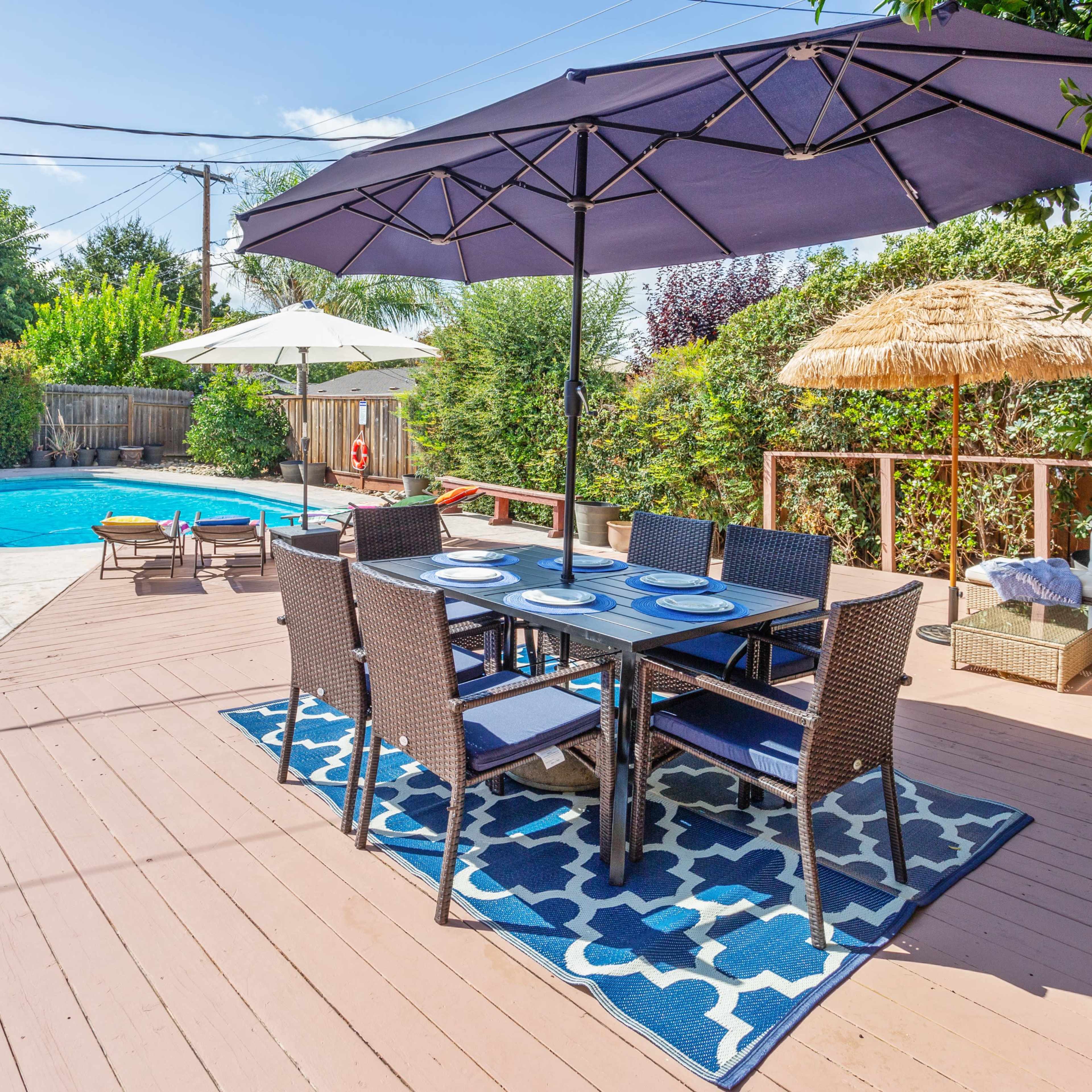 A backyard patio area with a dining table under a large umbrella, surrounded by a swimming pool and various seating arrangements.