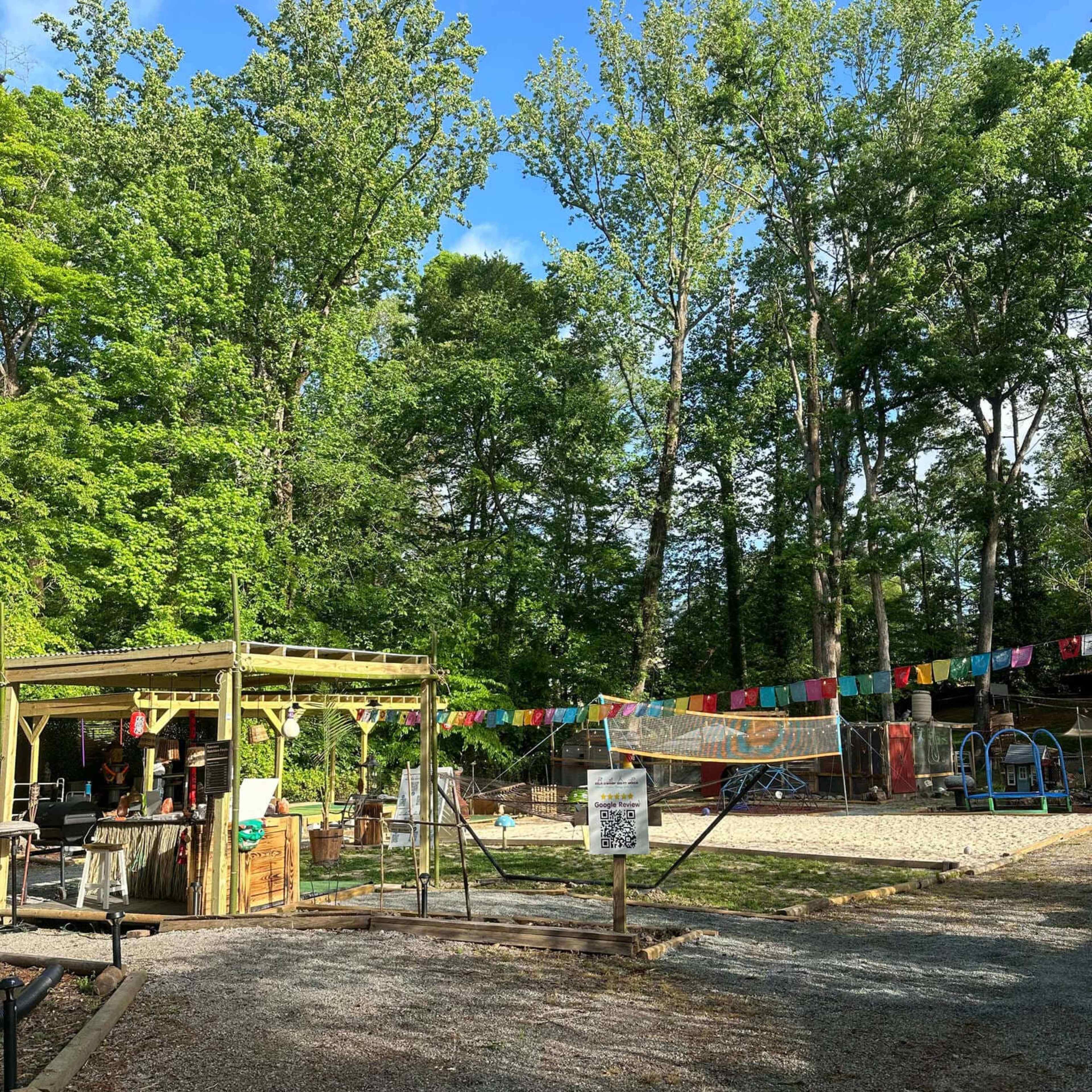 A gravel pathway leads to a gathering area surrounded by tall trees and colorful flags strung between them.