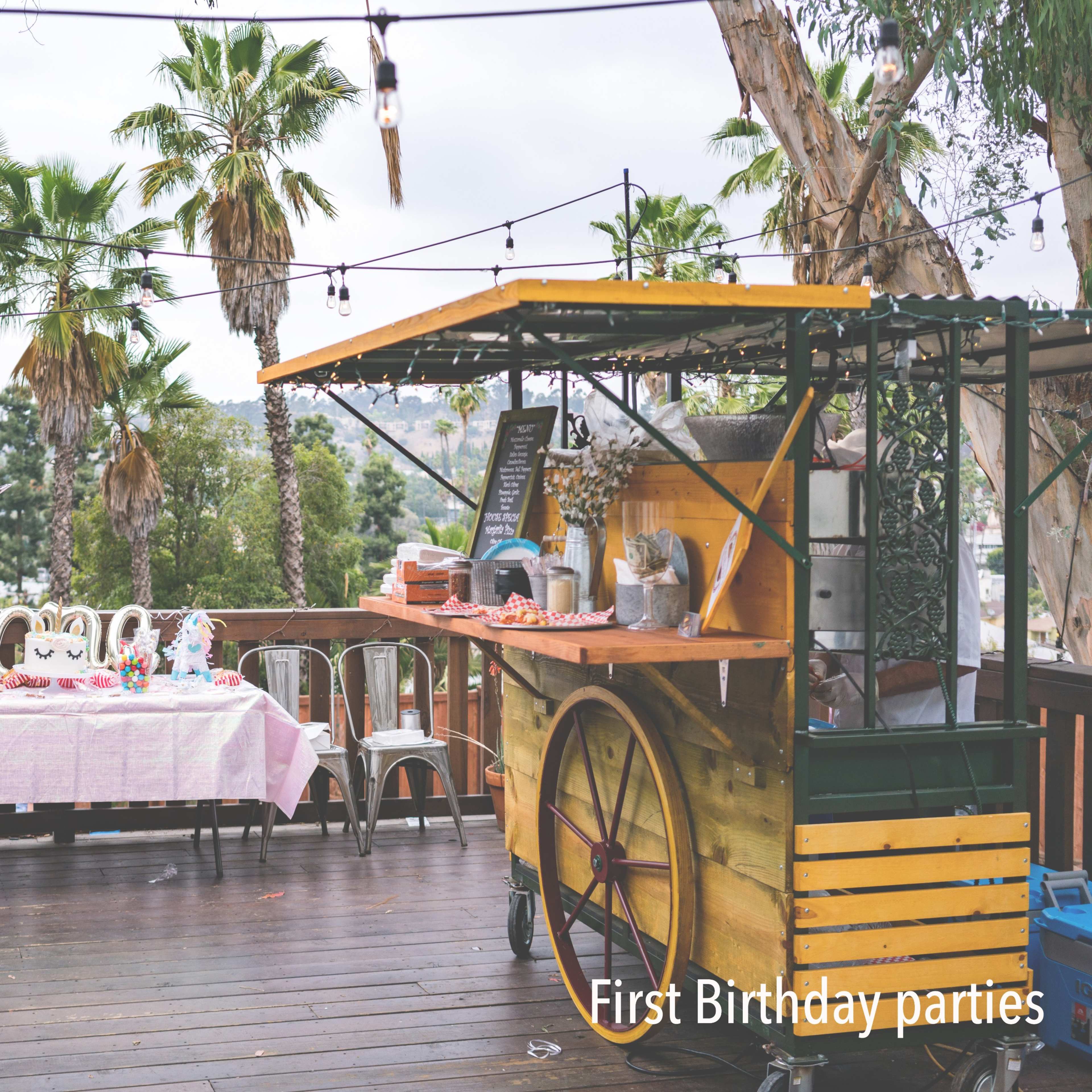 A wooden deck features a decorated table with a pink tablecloth and a food cart amidst palm trees in an outdoor setting.