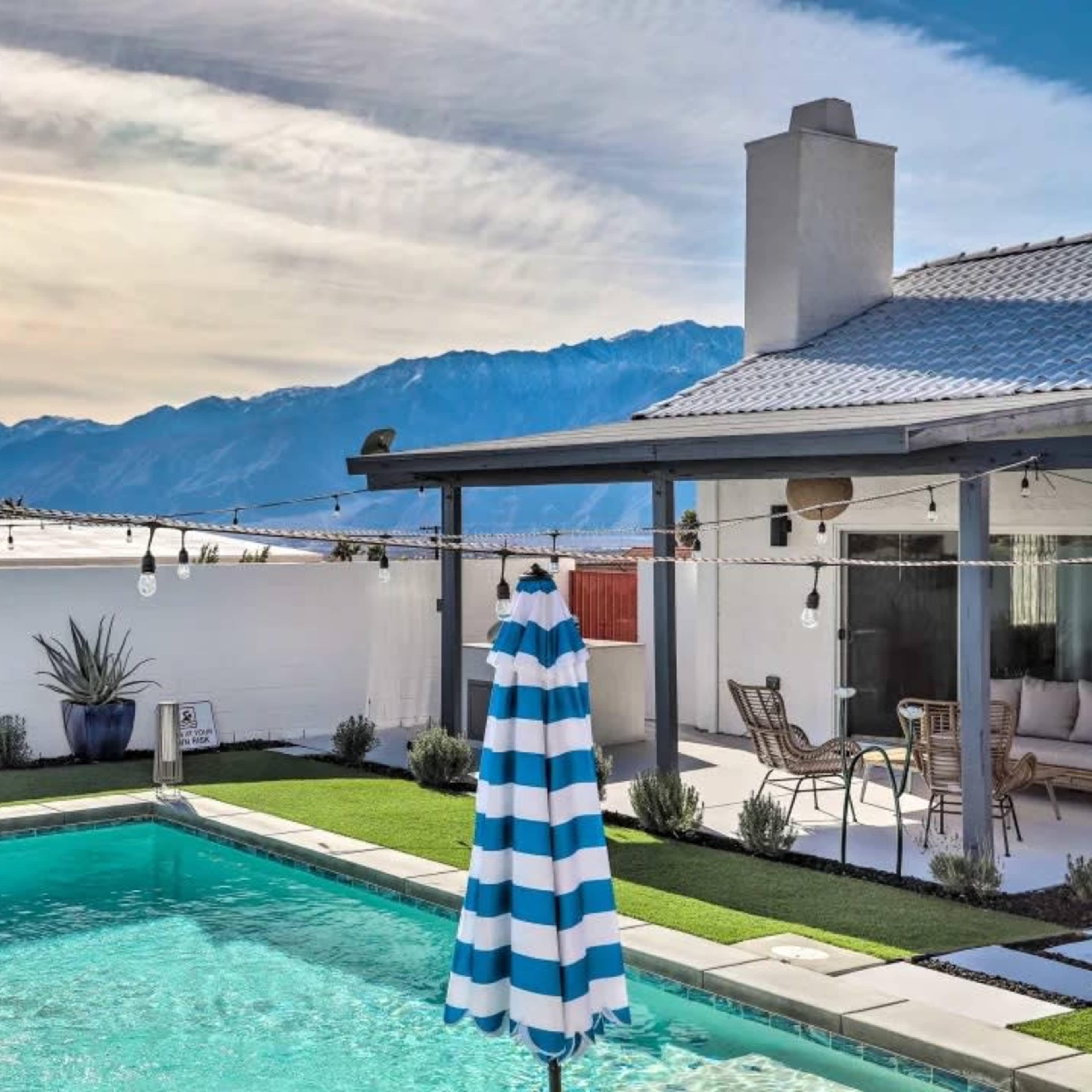 A swimming pool with a striped umbrella is situated beside a house, framed by mountains in the background.