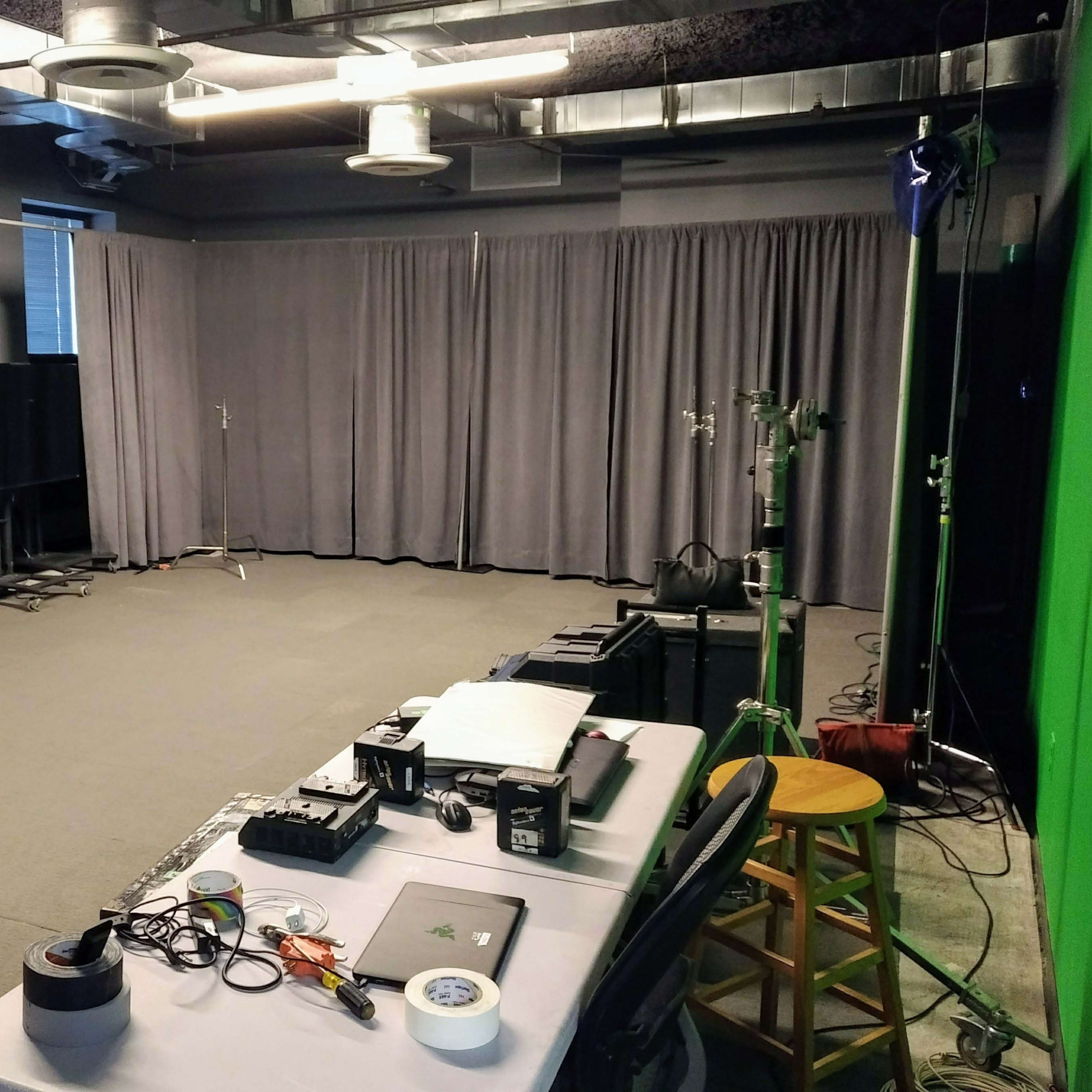 A production studio with grey curtains, a green screen, several cameras, and various technical equipment arranged on a table.