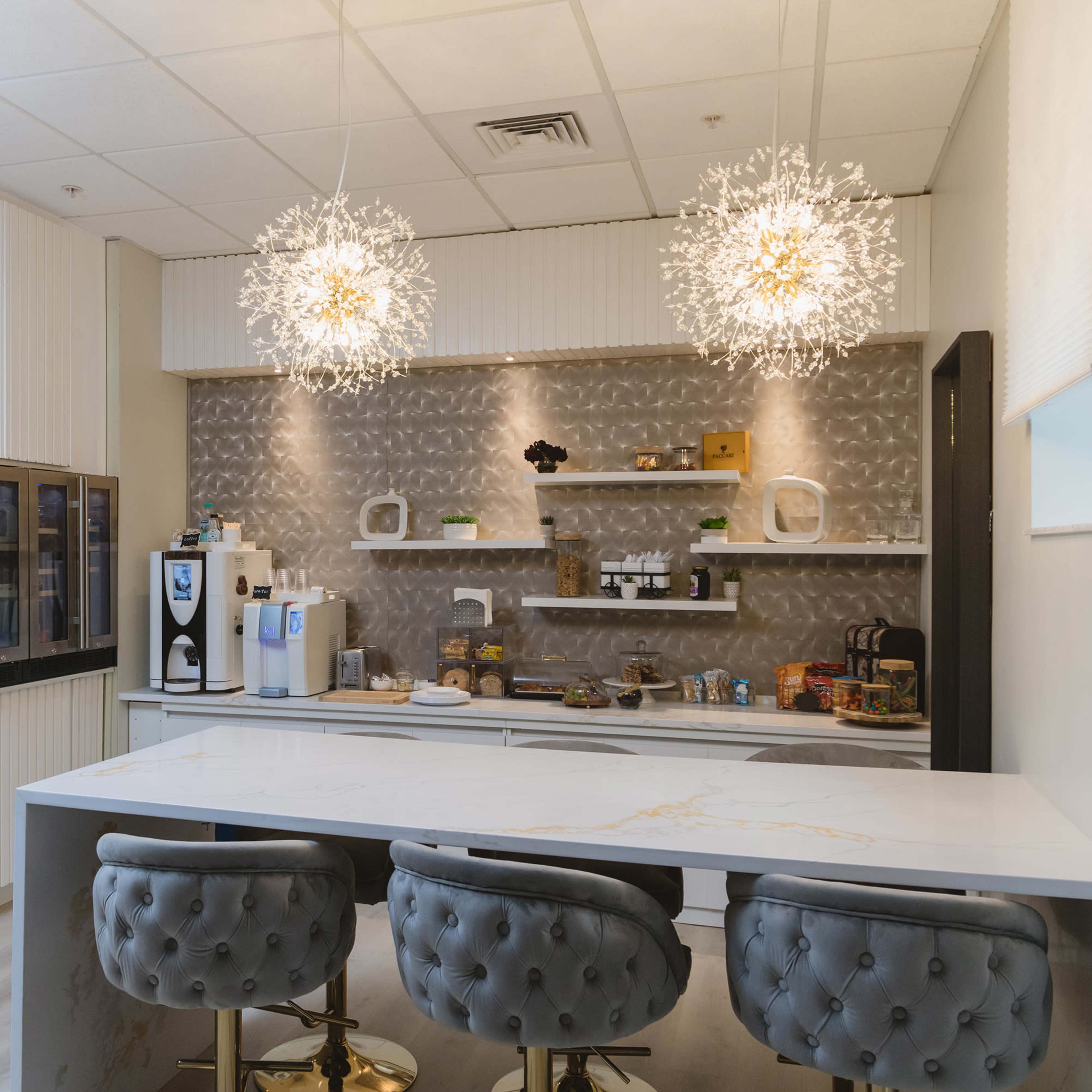 A modern kitchen with a white marble countertop, three tufted gray bar stools, and two decorative light fixtures hanging above.