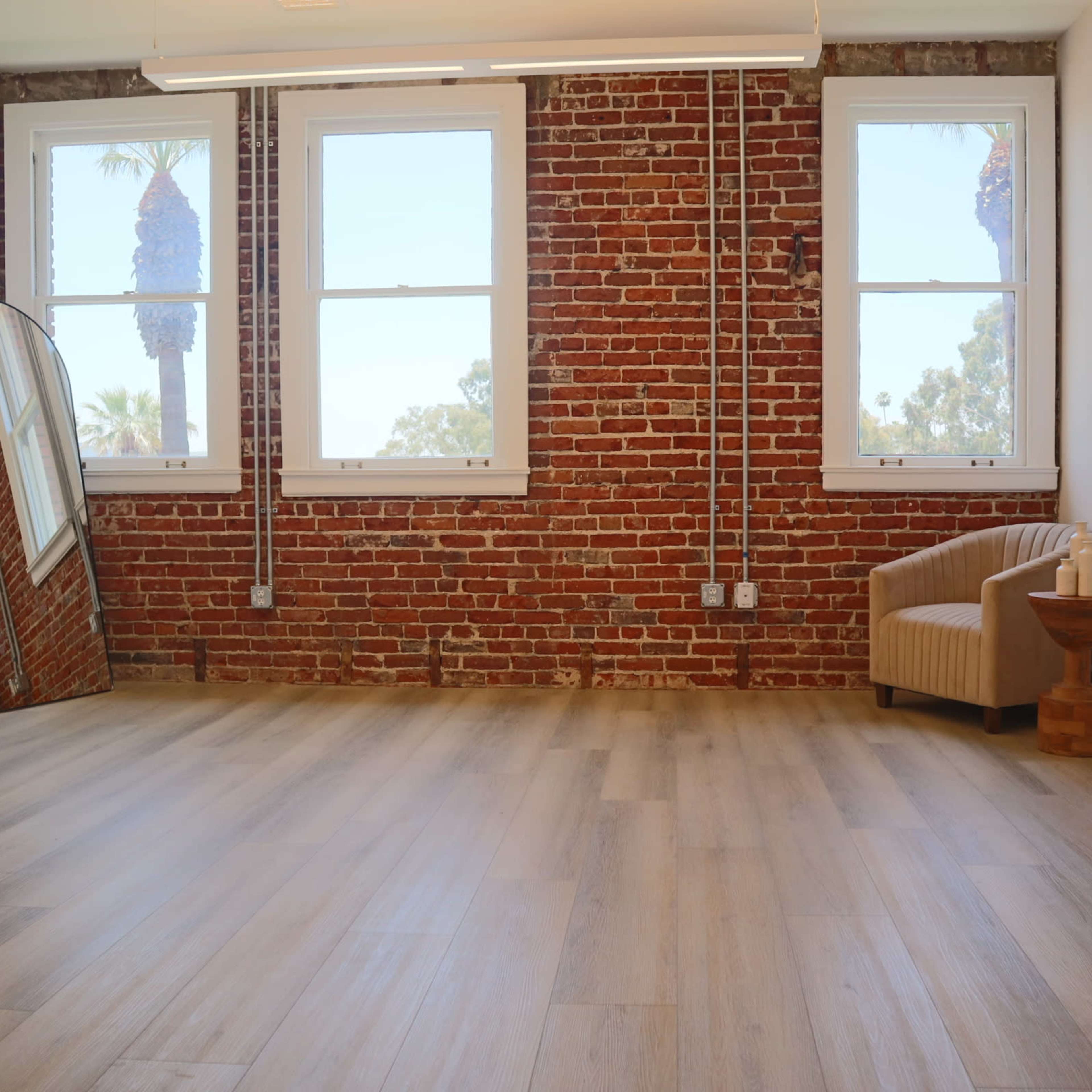 A spacious room with a brick wall, large windows, a curved mirror, a small wooden side table, and an upholstered chair.