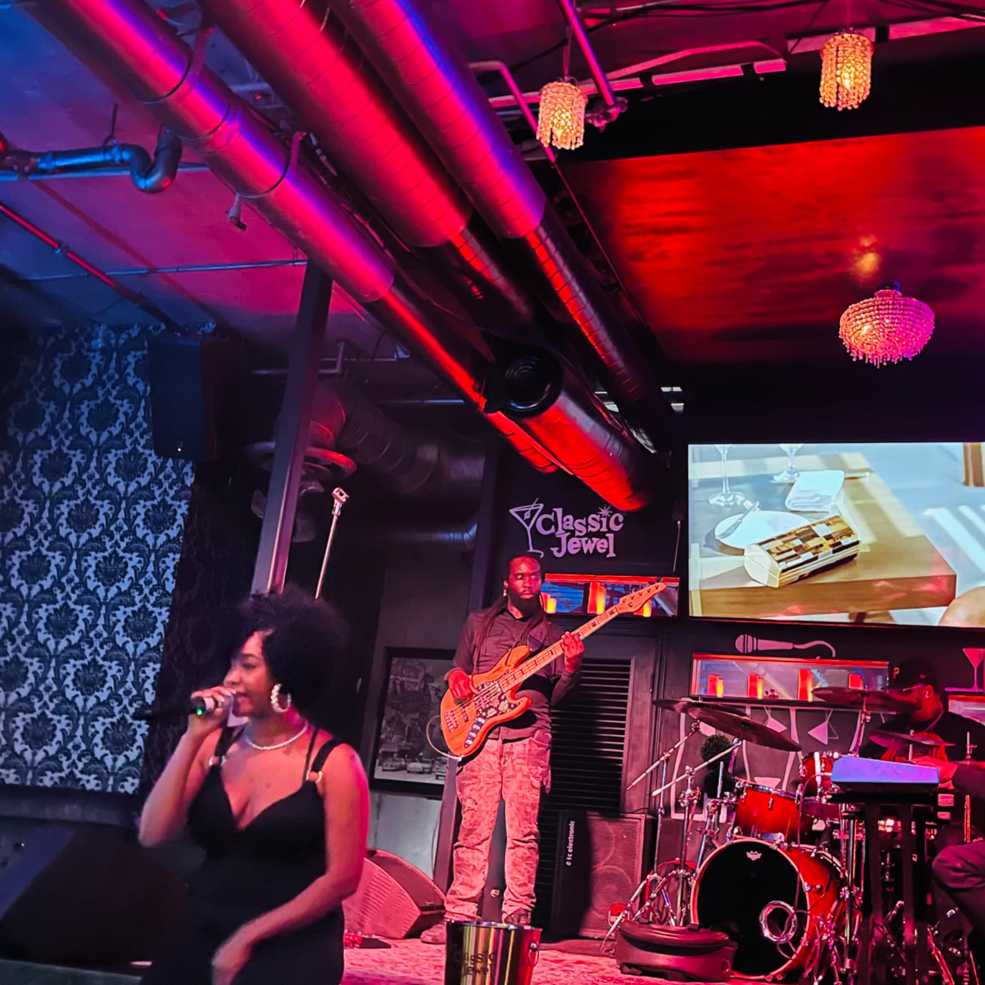 A woman sings on stage in front of a band while a video plays on a screen behind them in a dimly lit venue.