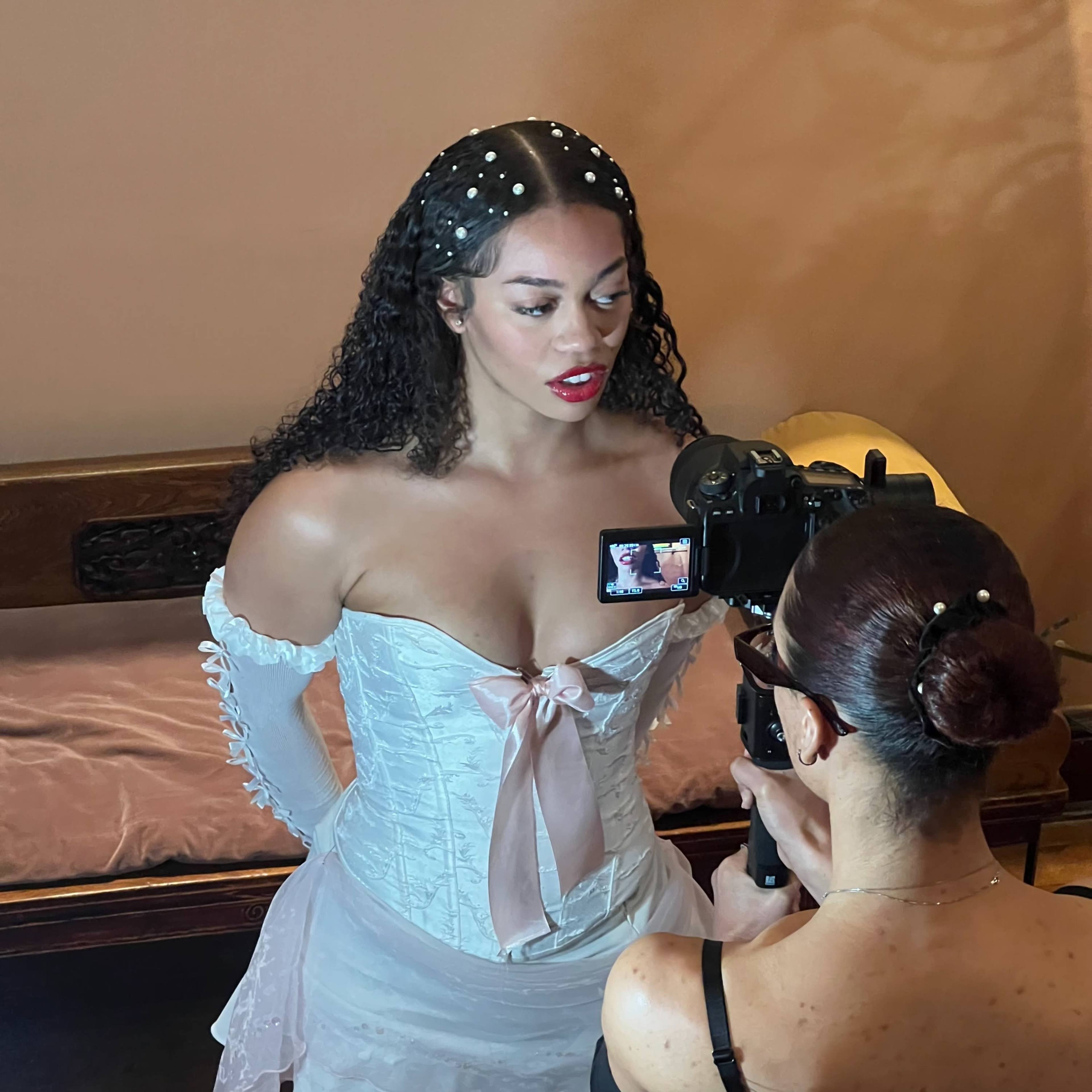 A woman in a white corset and dress is being filmed by another person holding a camera in a dimly lit room.