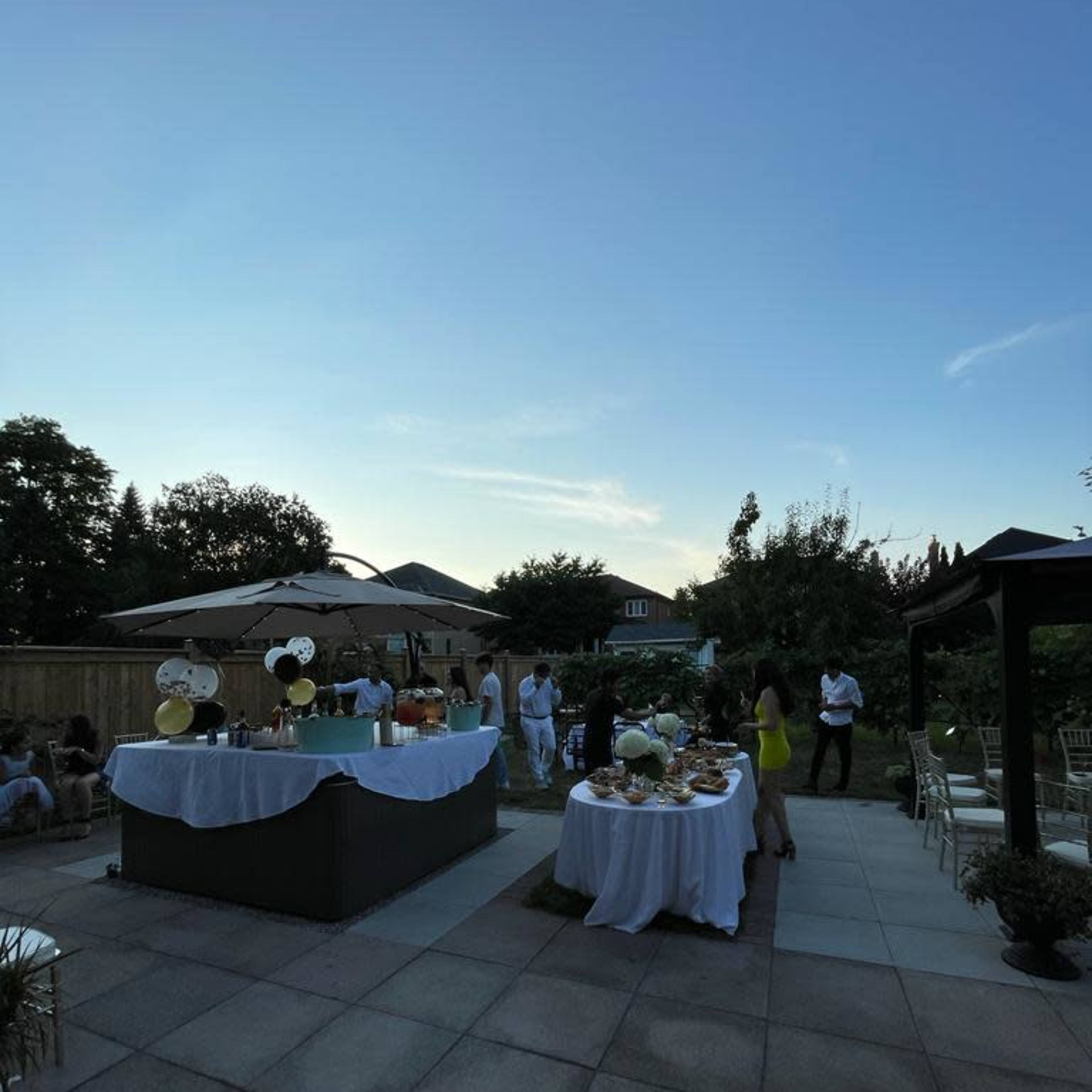 A backyard party features tables adorned with food and drinks, surrounded by guests enjoying the evening under a clear sky.