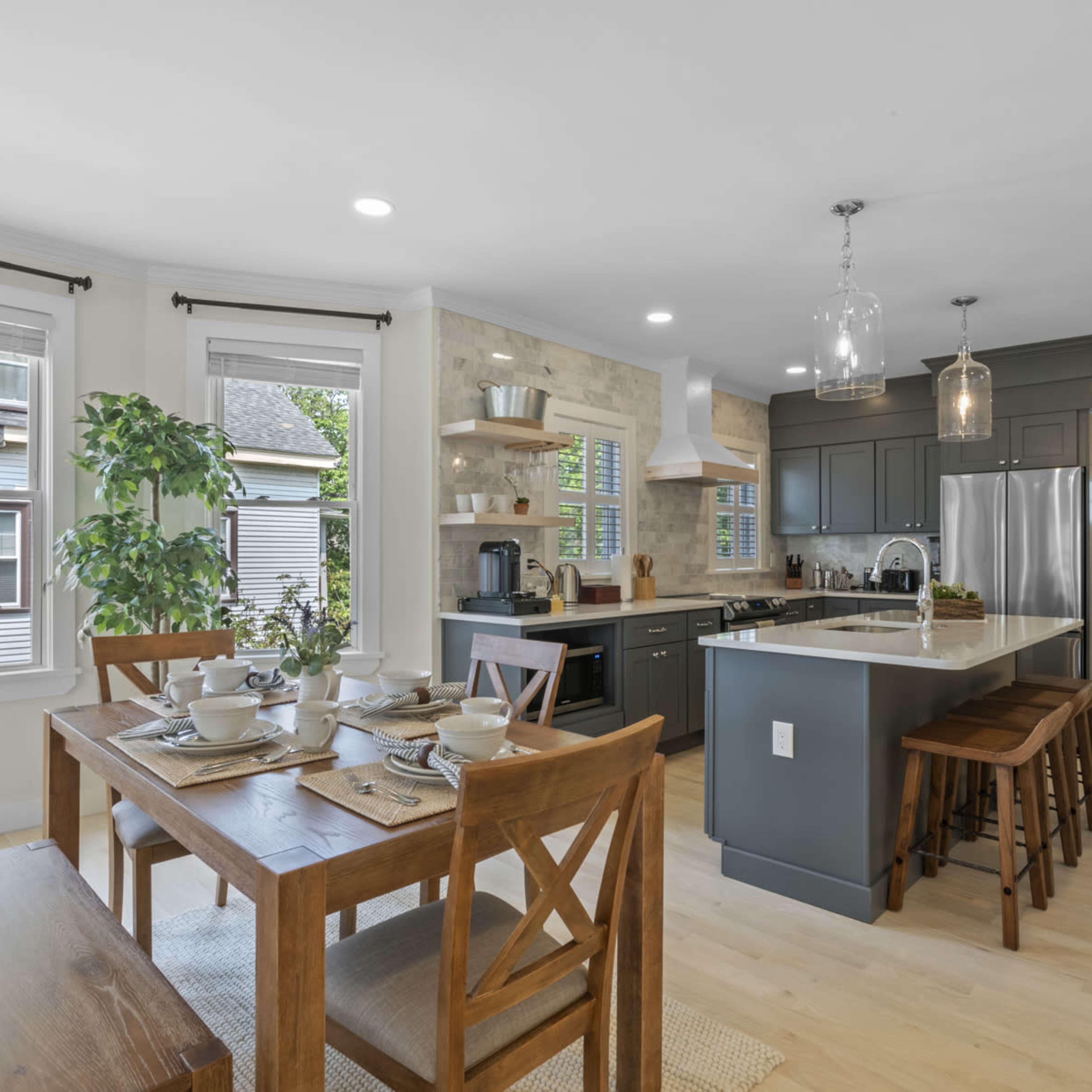 A modern kitchen features a central island with seating, a dining table set for four, and large windows allowing natural light to fill the space.