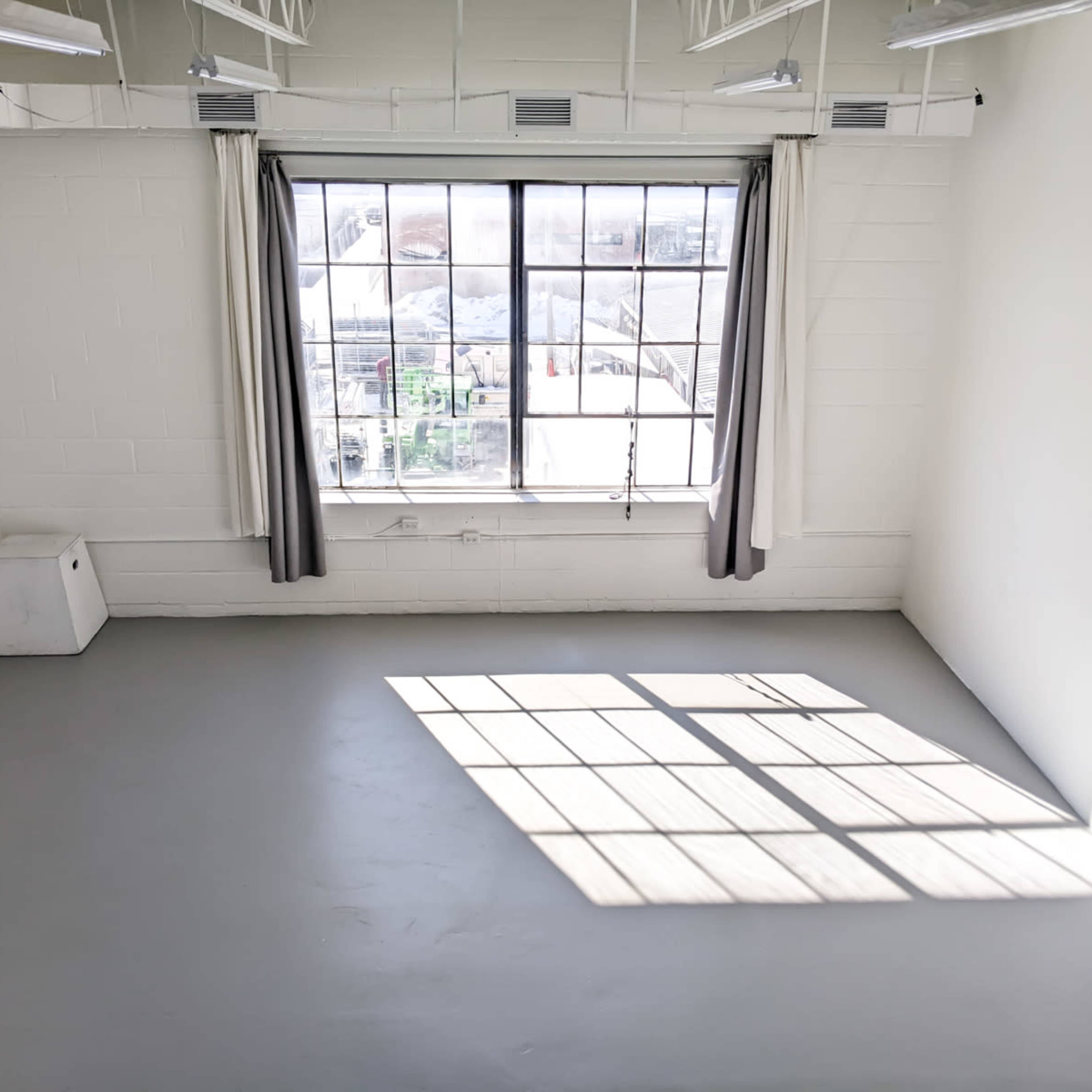 An empty room with gray flooring, a large window with curtains, and sunlight casting a rectangular shadow on the floor.