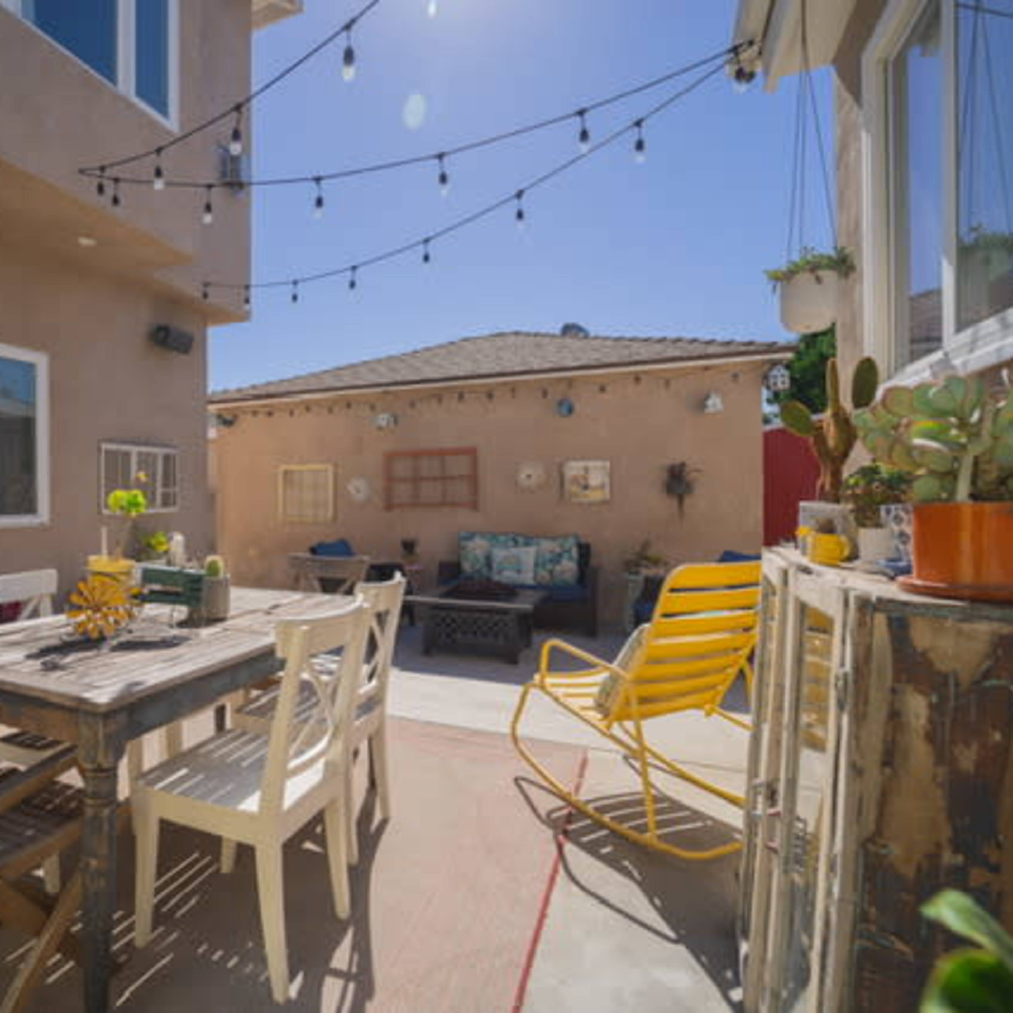 A sunny outdoor patio with a wooden dining table surrounded by chairs, string lights above, and potted plants along the walls.