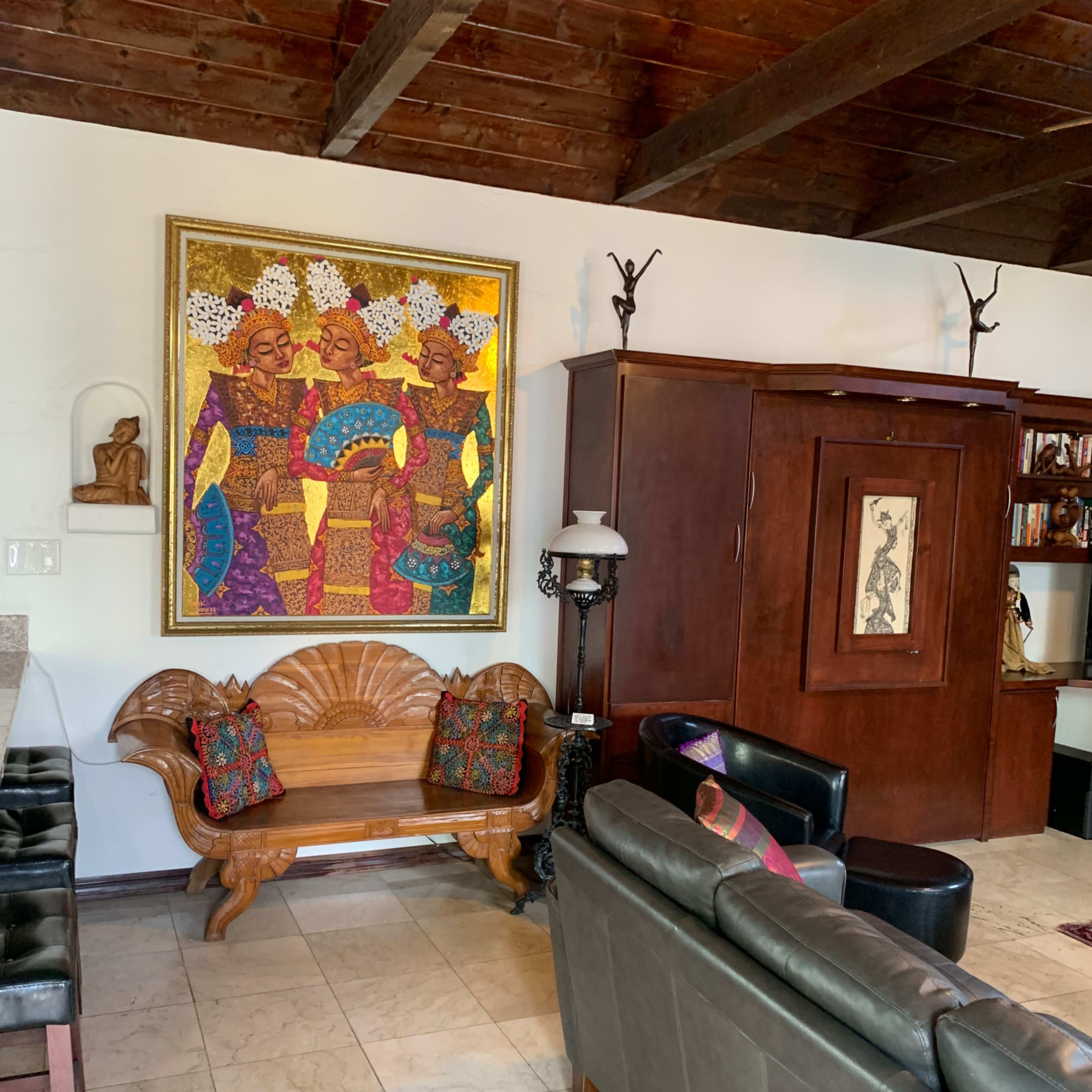 A living room with a wooden bench, a dark leather sofa, and a large colorful painting featuring three figures on the wall.