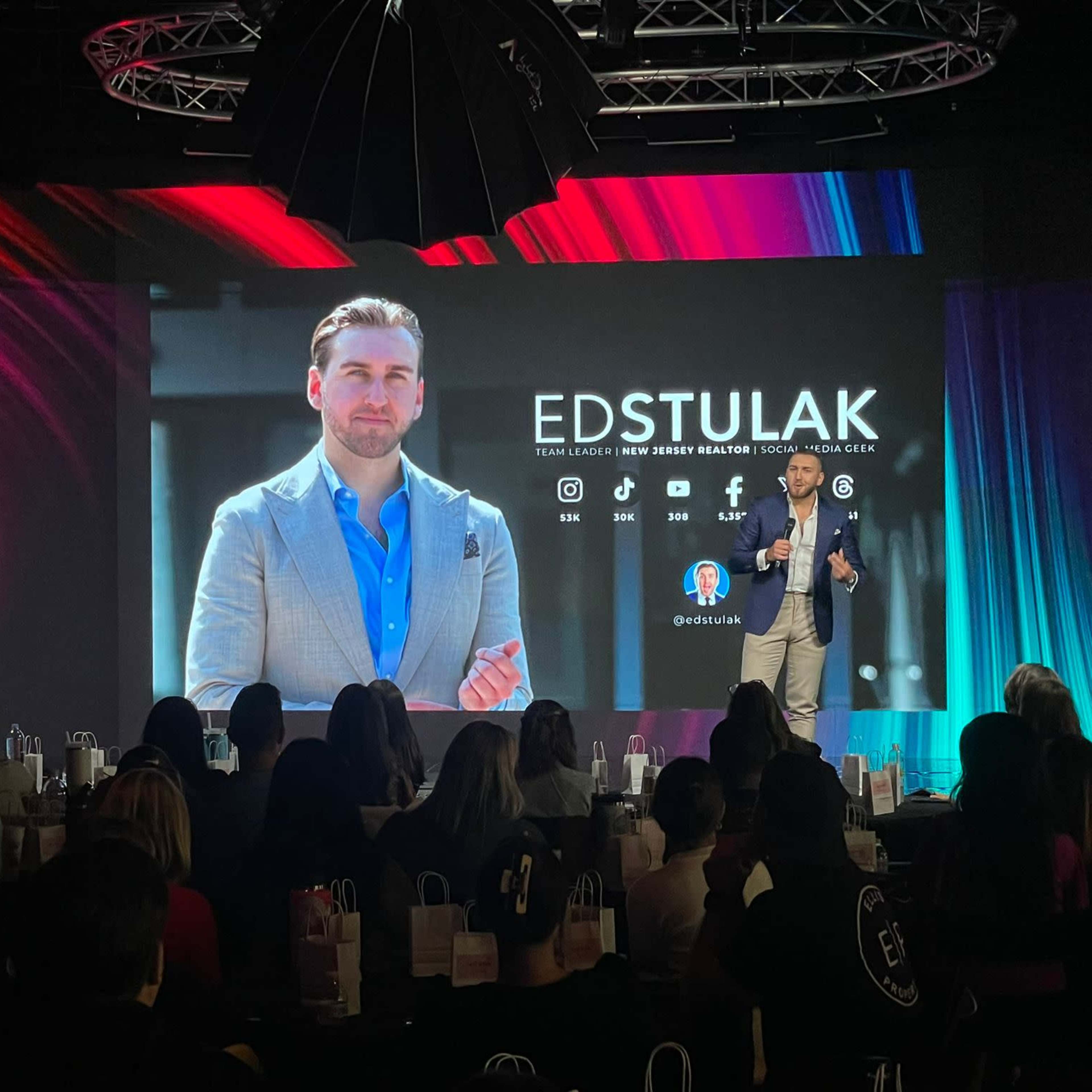 A speaker presents on stage to an audience while a large screen displays a portrait and information about Ed Stulak.