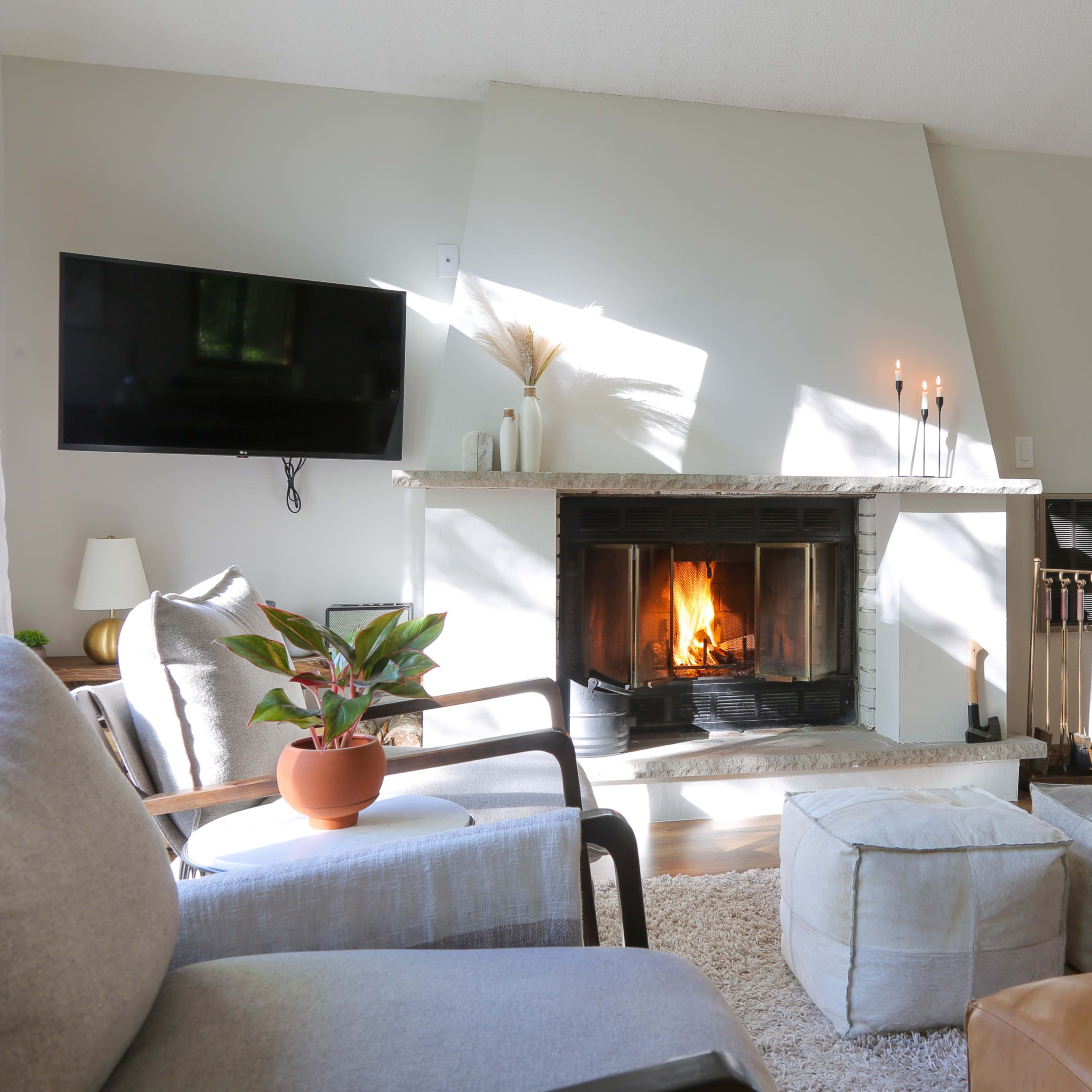 A cozy living room features a stone fireplace with a lit fire, a television mounted on the wall, and comfortable seating arrangements, including a gray armchair and a brown leather couch.