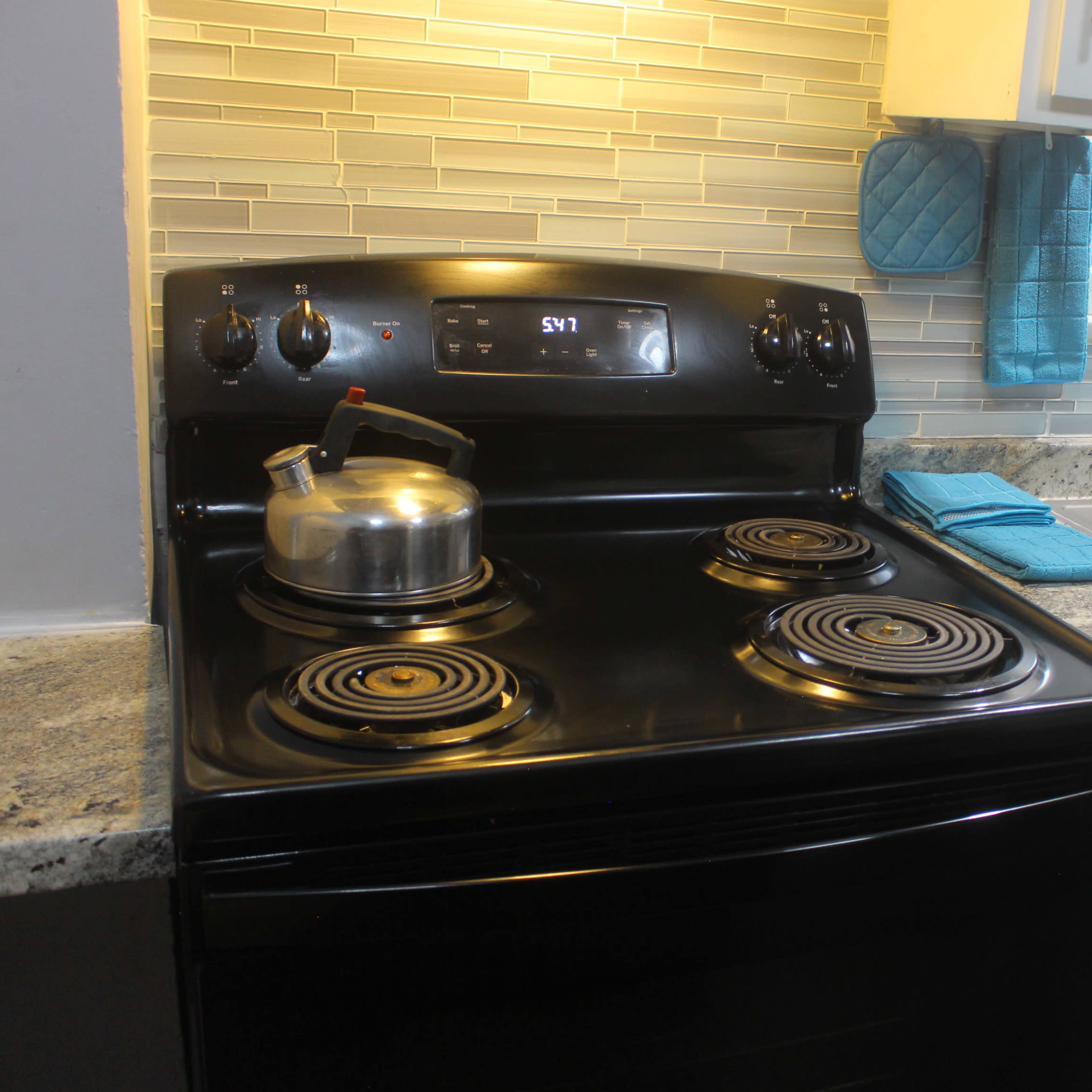 A black stove with a kettle on one burner, adjacent to a kitchen sink and countertop with a light blue dish towel.