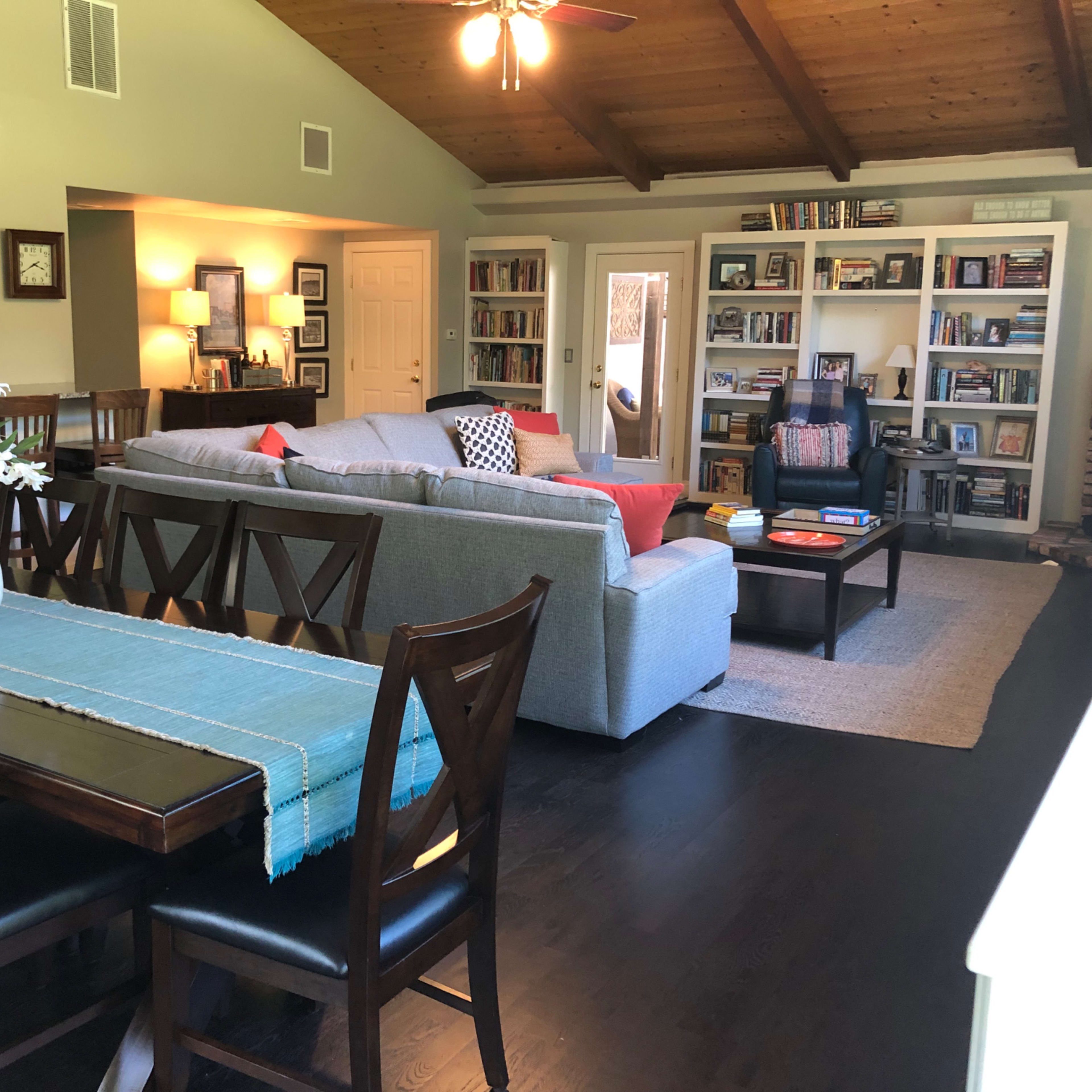 A spacious living area featuring a gray sectional sofa, a coffee table, bookshelves filled with books, and a dining table with chairs, all under a wooden ceiling.