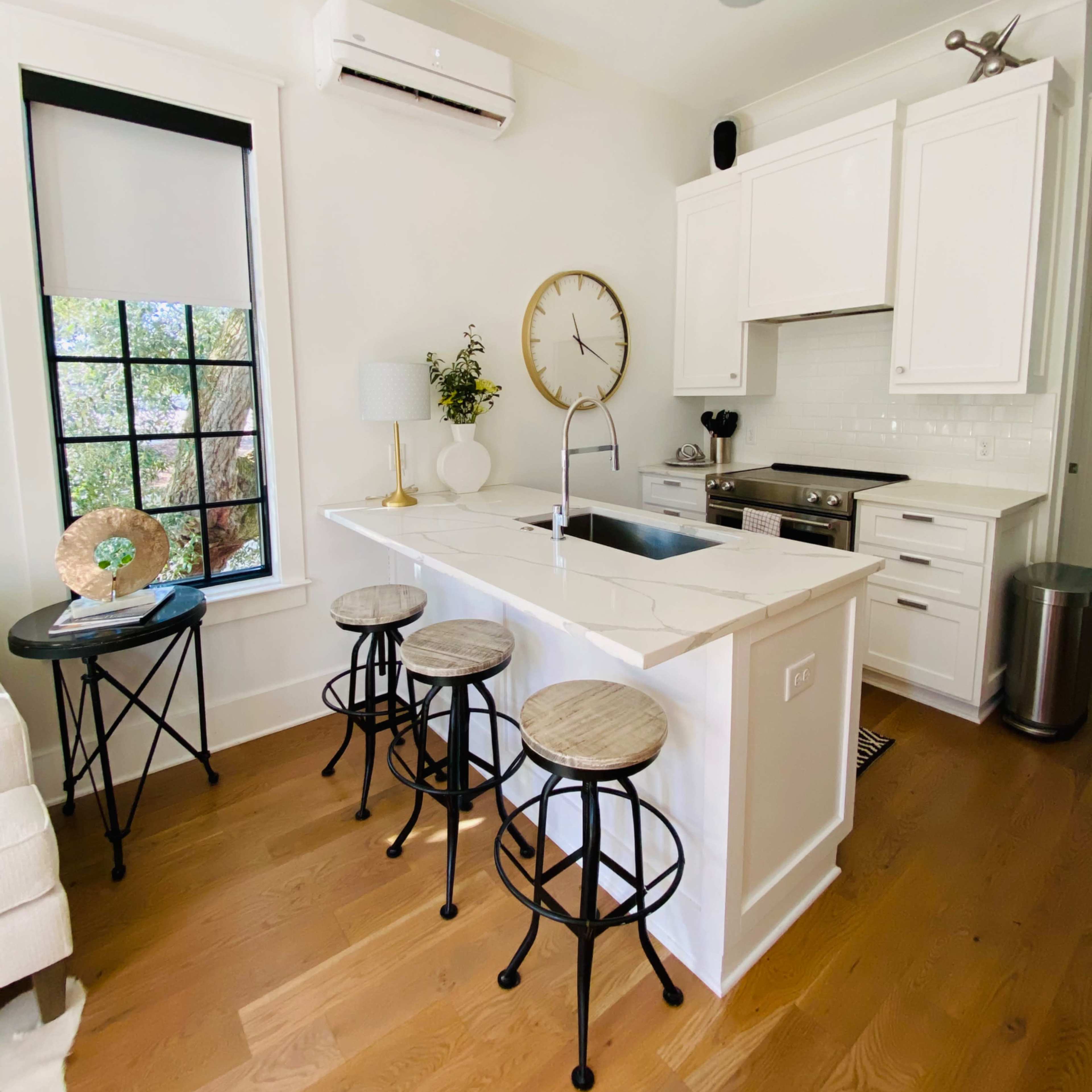 A modern kitchen features a white island with three wooden bar stools, stainless steel appliances, and a clock on the wall.