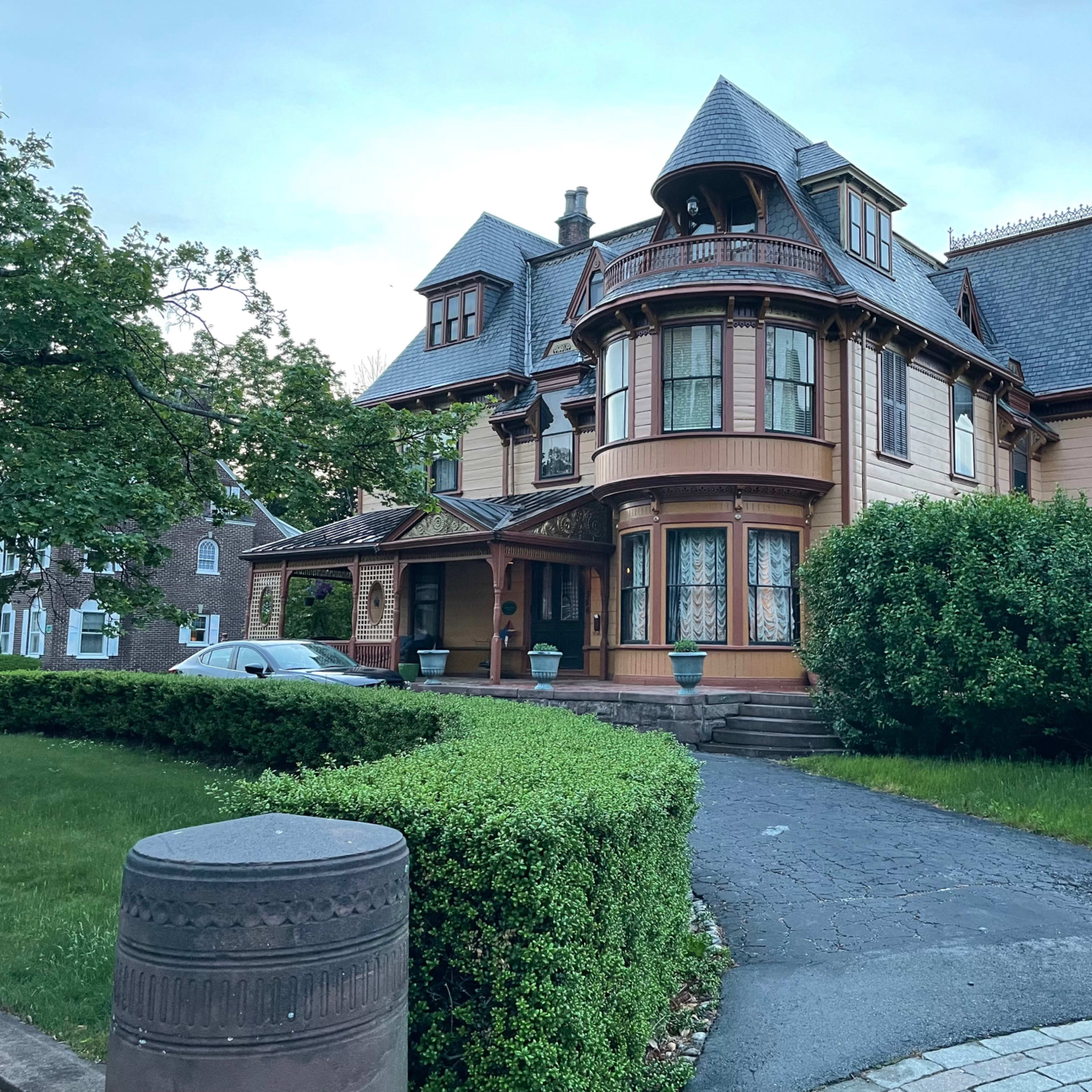 A large Victorian-style house with a rounded turret and intricate woodwork, surrounded by green shrubbery and a manicured lawn.