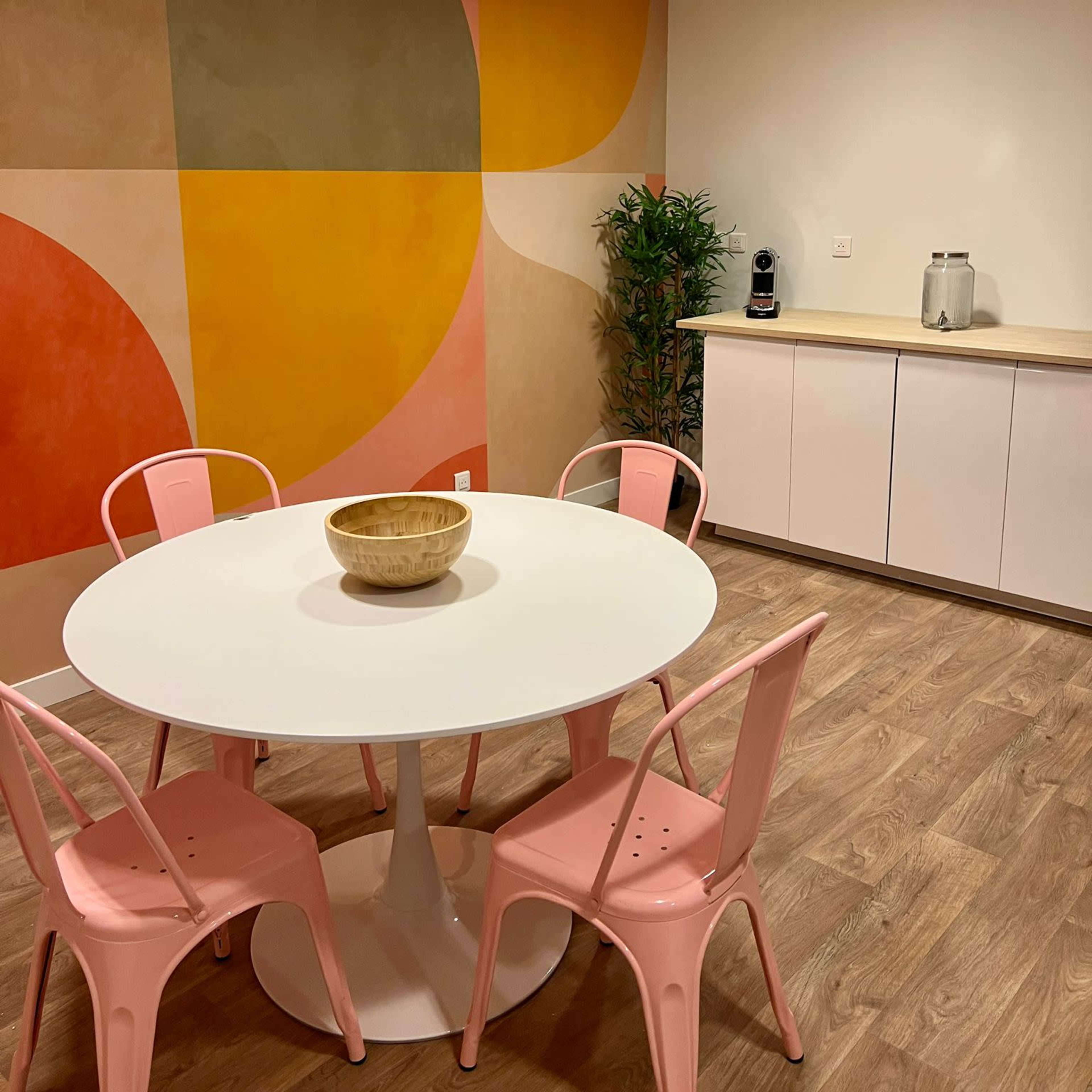 A minimalist dining area with a round white table at the center, surrounded by pink chairs, against a backdrop of colorful geometric wall art.