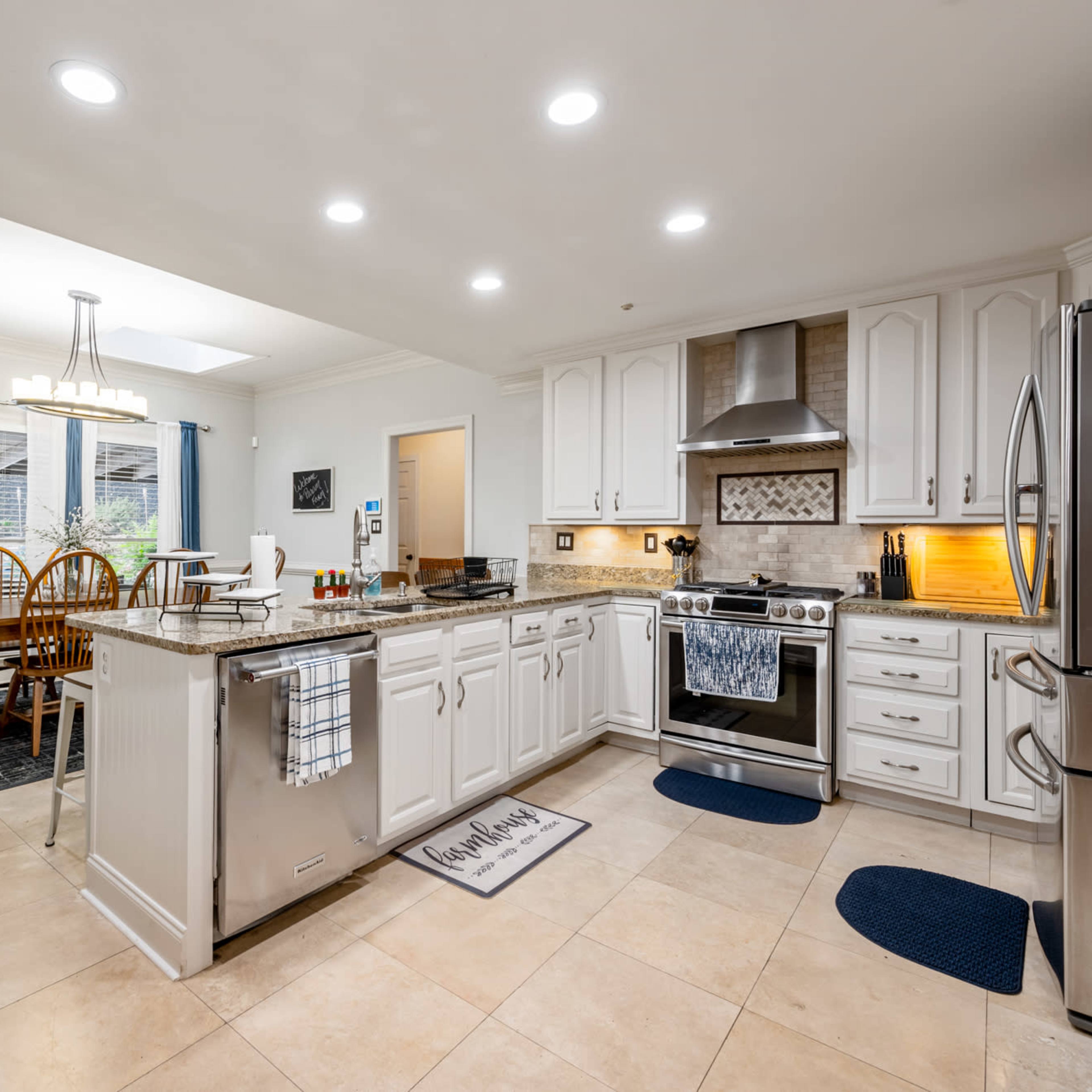 A kitchen with white cabinetry, a stainless steel stove, a dining table with chairs, and large windows letting in natural light.