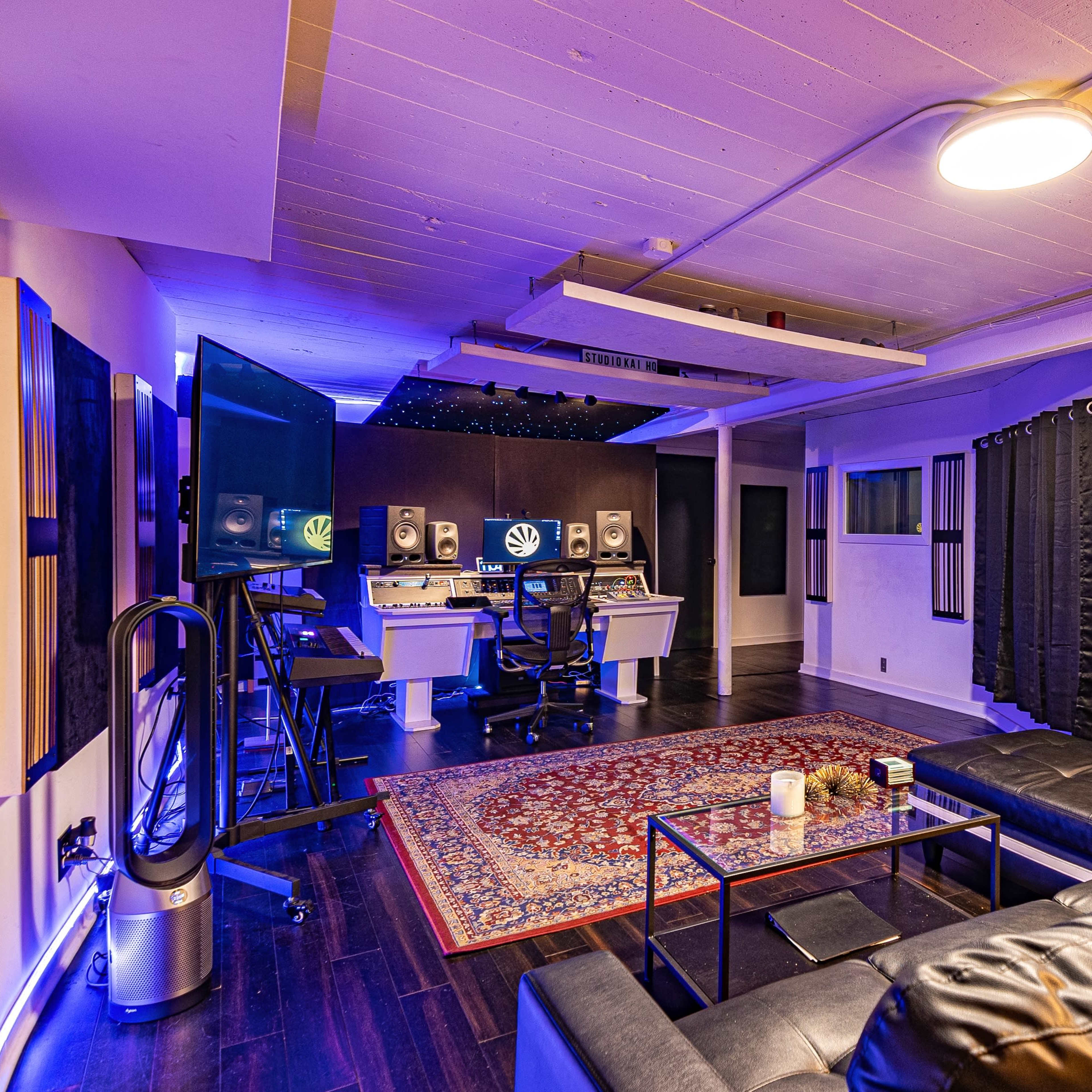 A well-equipped music studio featuring two computer stations, black acoustic panels, a seating area with a couch, and ambient lighting.