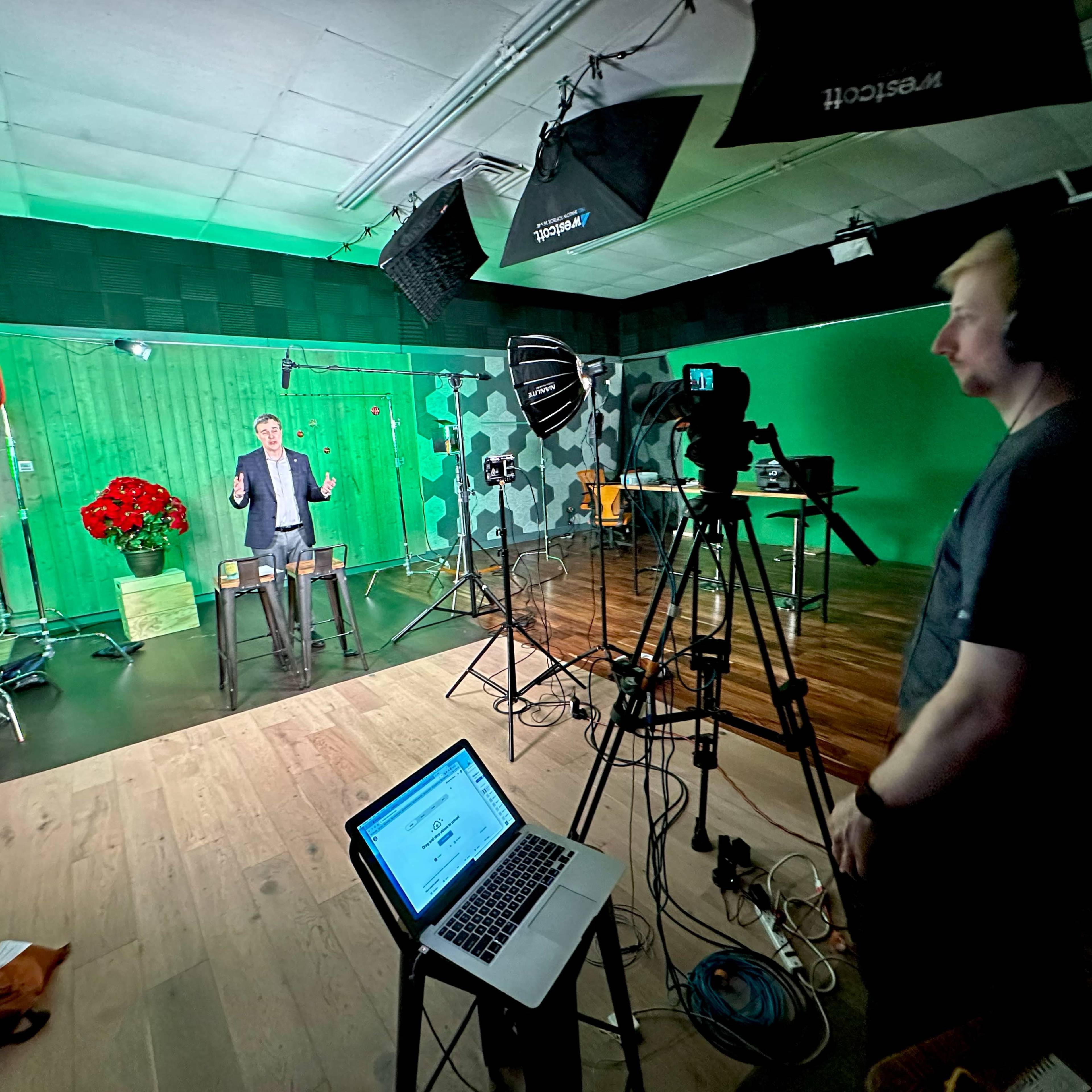 A man stands on a set with green screens and studio lights, while a crew member operates a camera and monitors equipment.