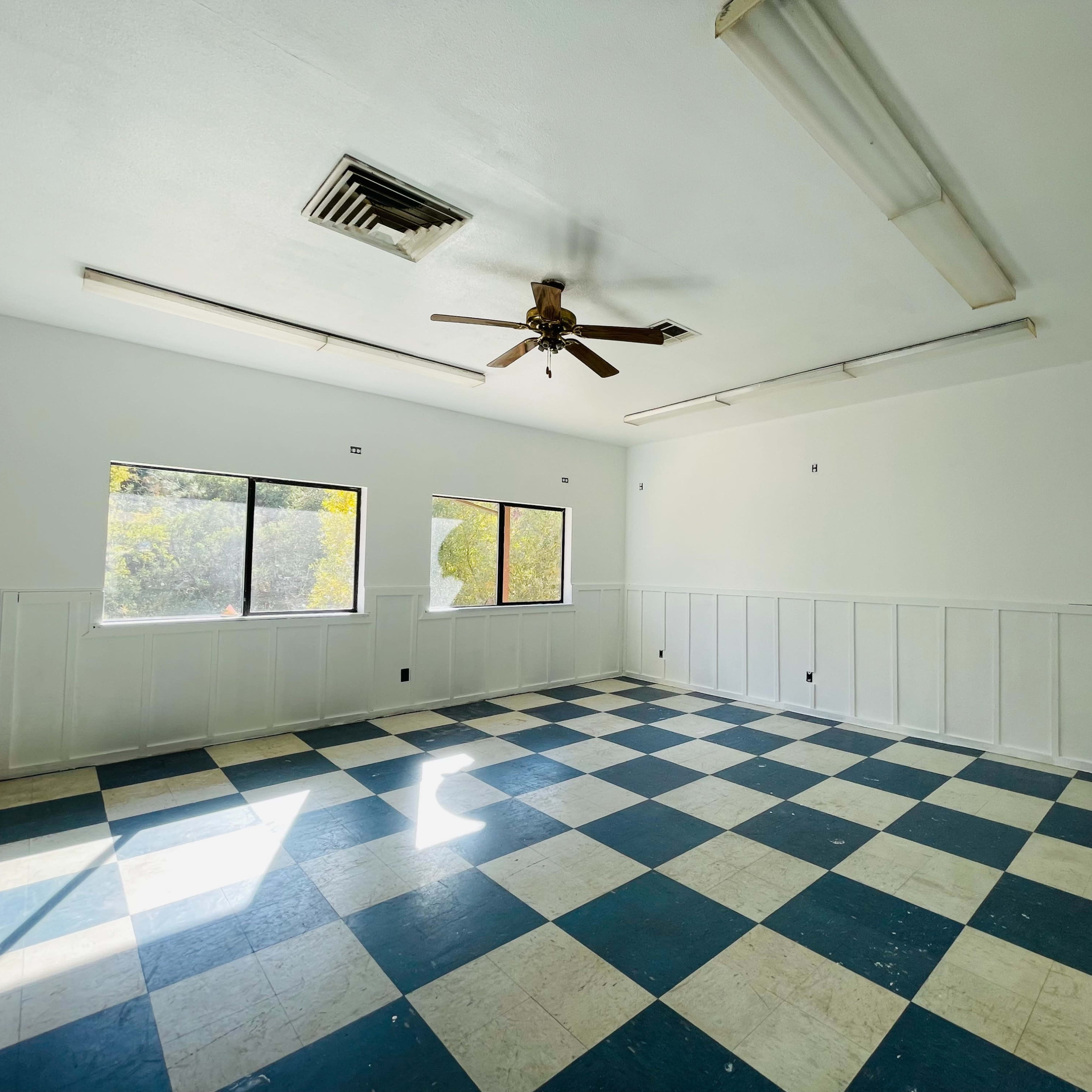 An empty room with a checkered blue and white floor, two windows, and a ceiling fan.