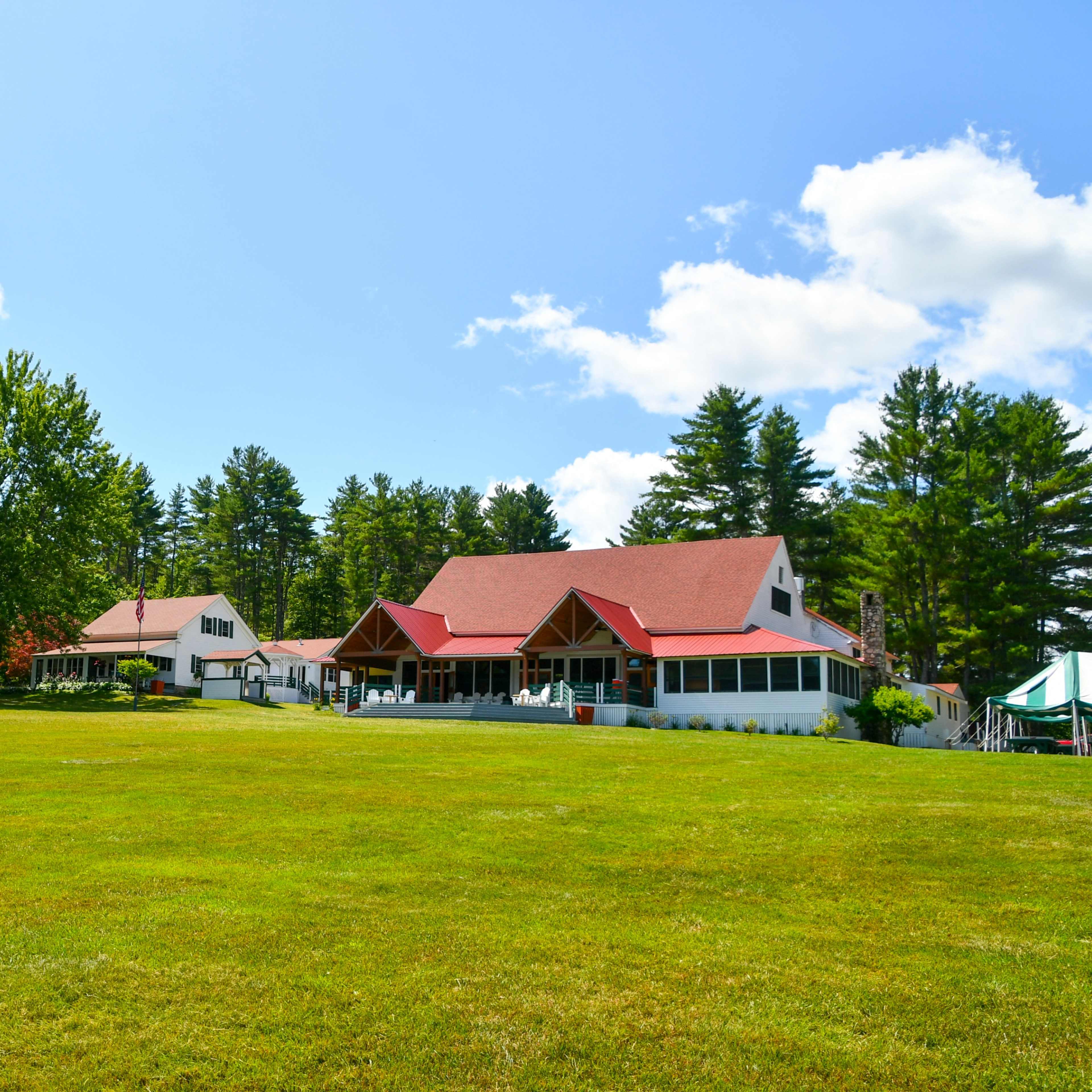 A large, open grassy area features several buildings with red roofs surrounded by tall trees under a clear blue sky.