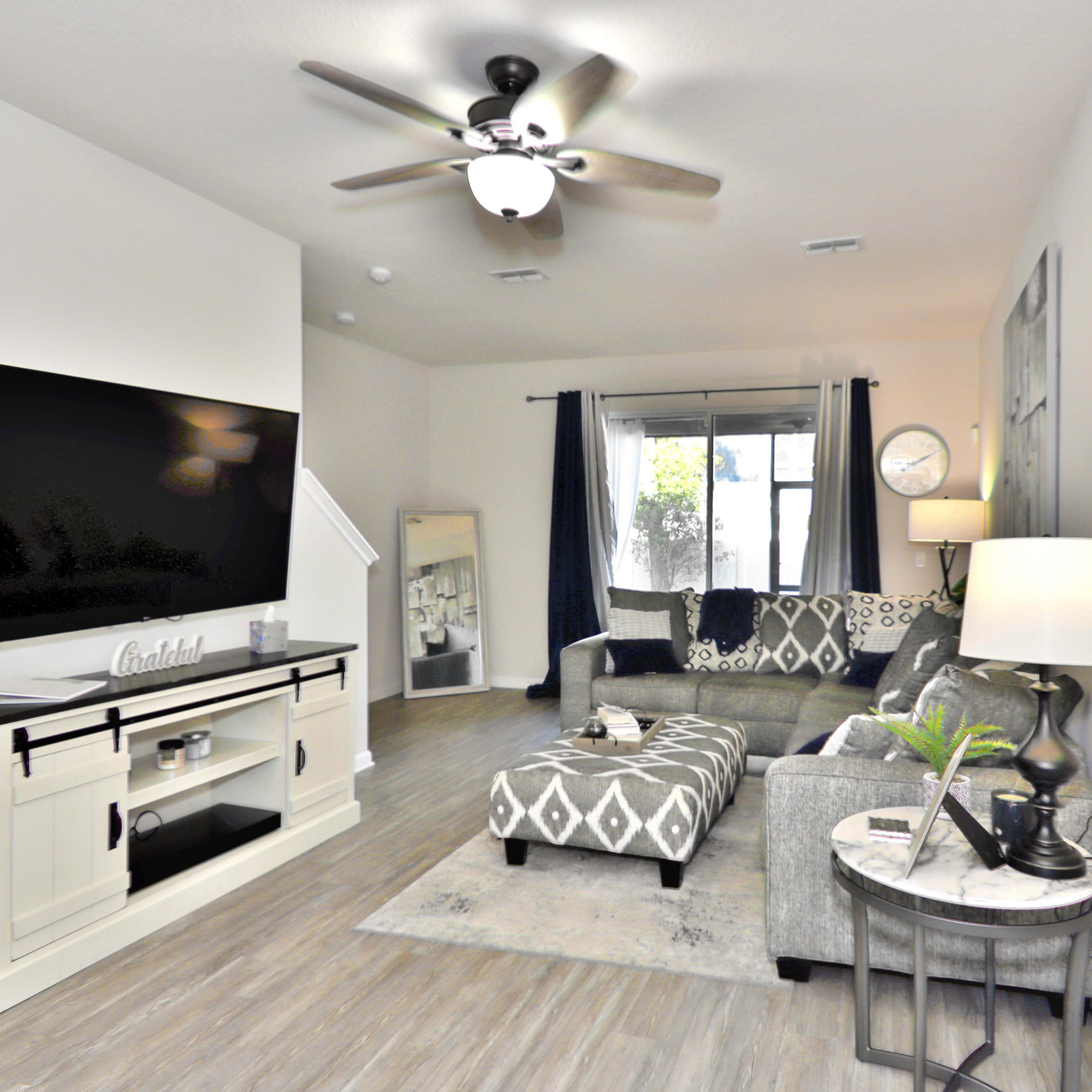 The living room features a sectional couch, a large flat-screen TV mounted on the wall, and a lamp beside a round table, creating a cozy and modern space.