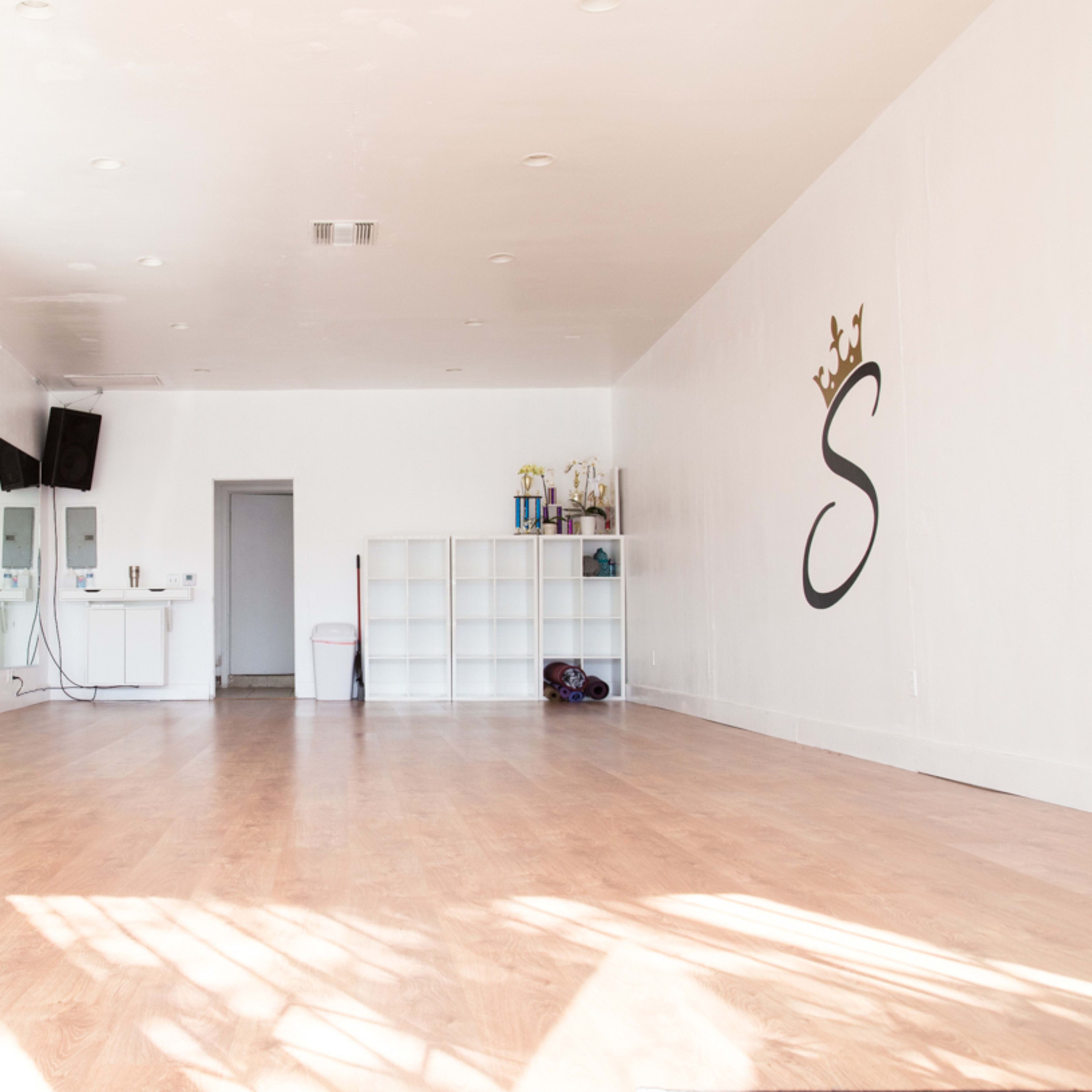 A spacious dance studio with a mirrored wall, wooden floor, and a large wall-mounted letter "S."