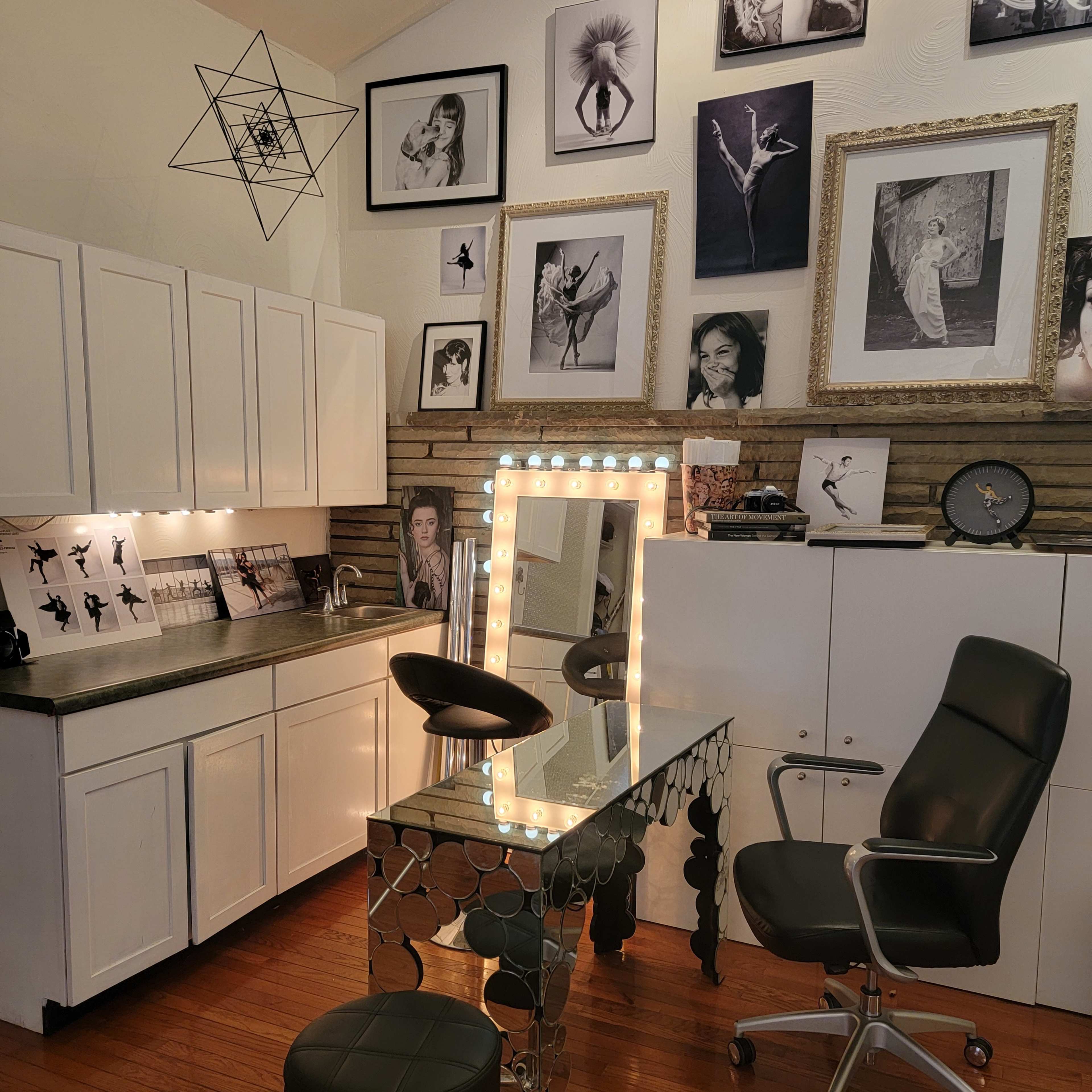 A brightly lit makeup station with a large mirror surrounded by lights, set against a wall adorned with black-and-white photographs and a sleek, modern design in a well-organized room.