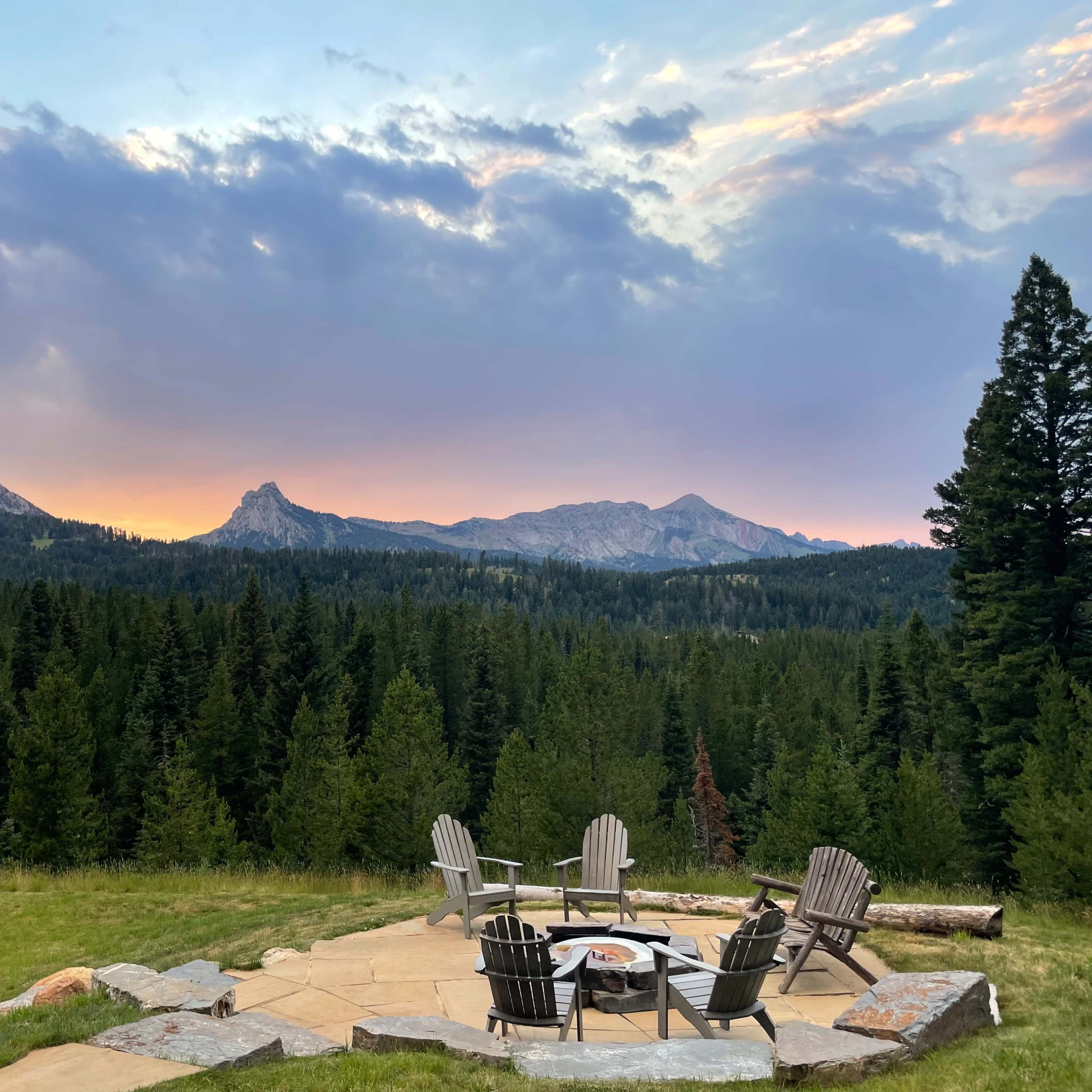 A stone patio with wooden chairs surrounds a fire pit, set against a backdrop of mountains and a forested landscape under a colorful sky.