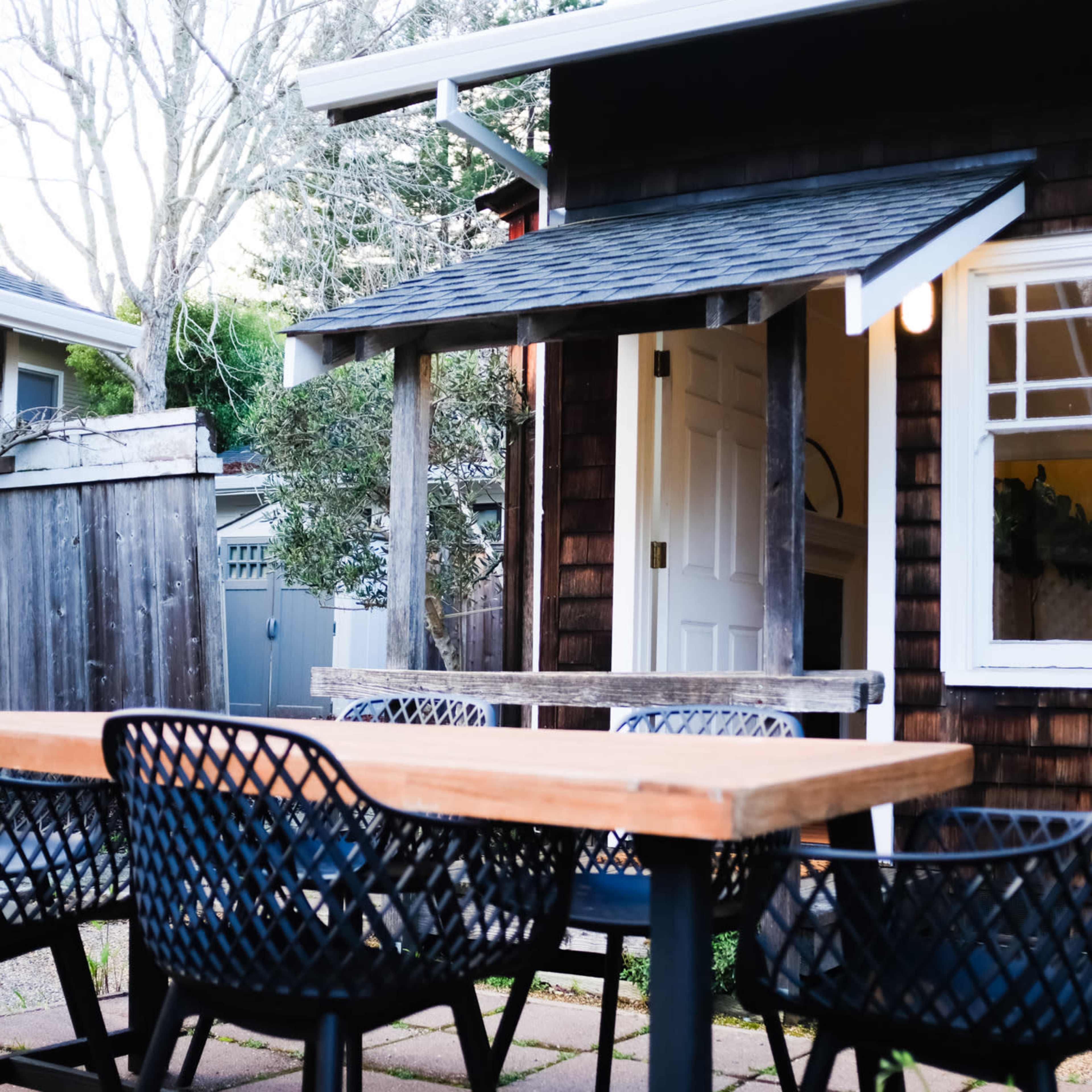 A wooden table with black woven chairs is set in a patio area beside a small house with a chimney and an open door.