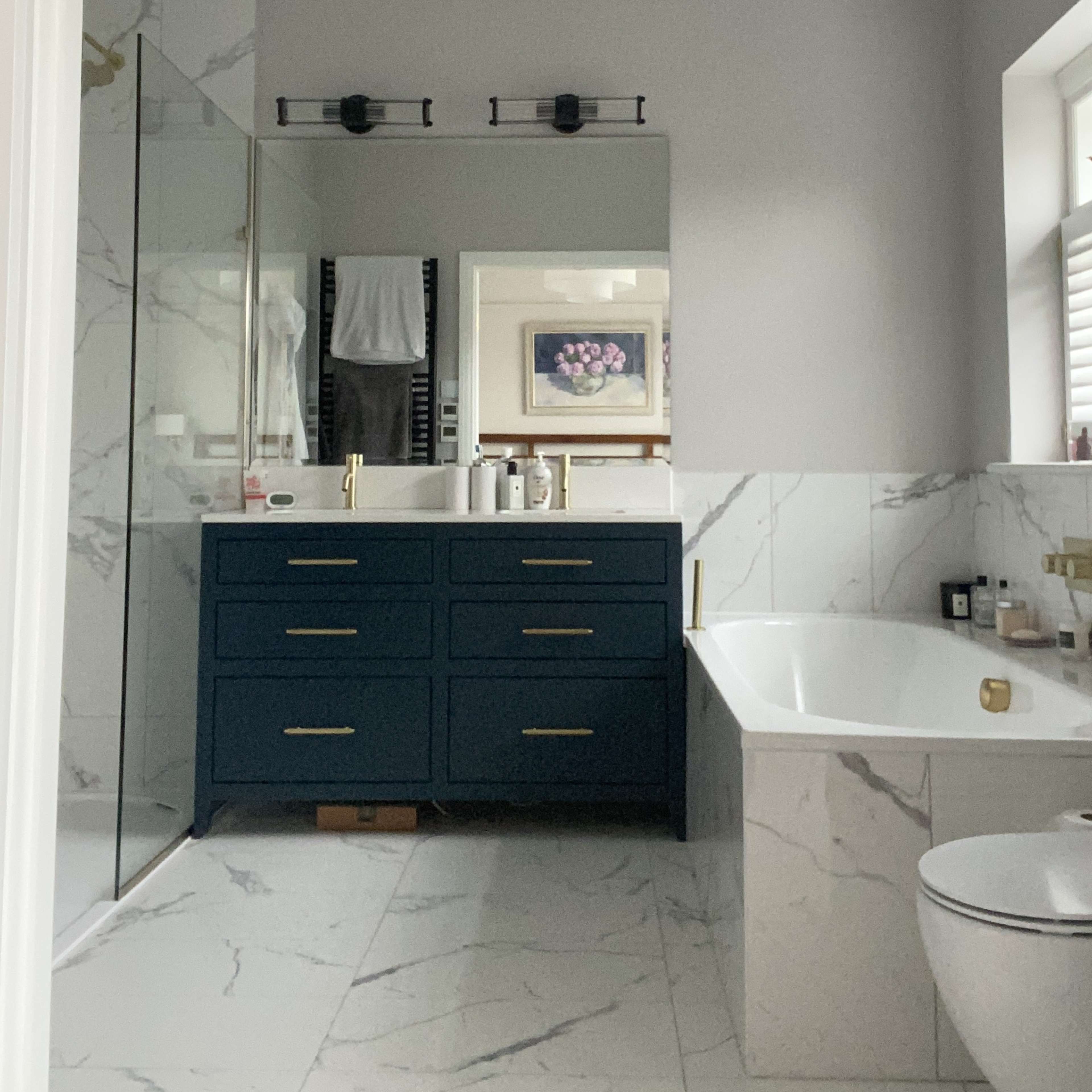 A modern bathroom featuring a dark blue vanity with gold fixtures, a large mirror, a glass shower enclosure, a freestanding bathtub, and marble-patterned tiles.