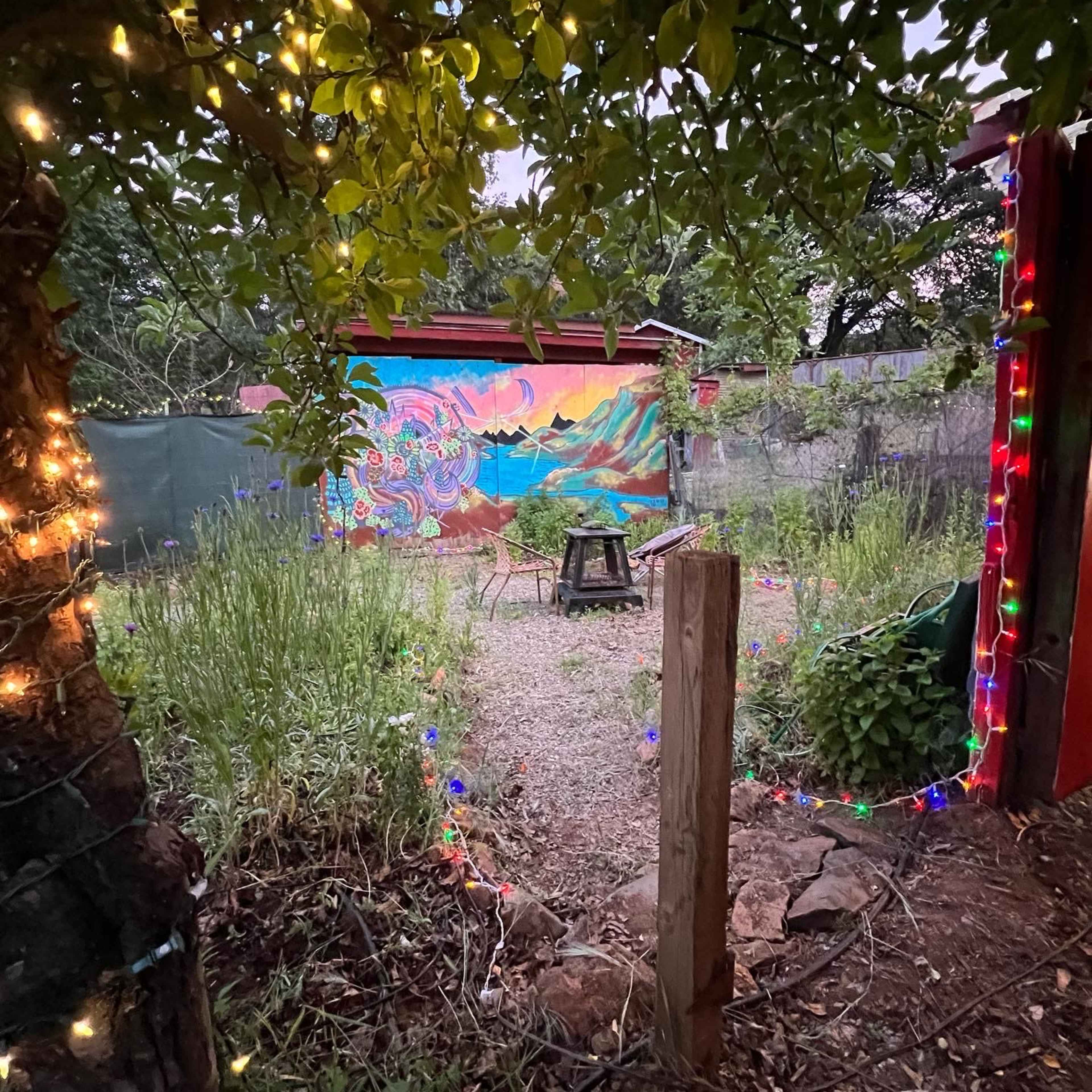 A vibrant mural decorates the back wall of a garden surrounded by plants and lined with colorful string lights.