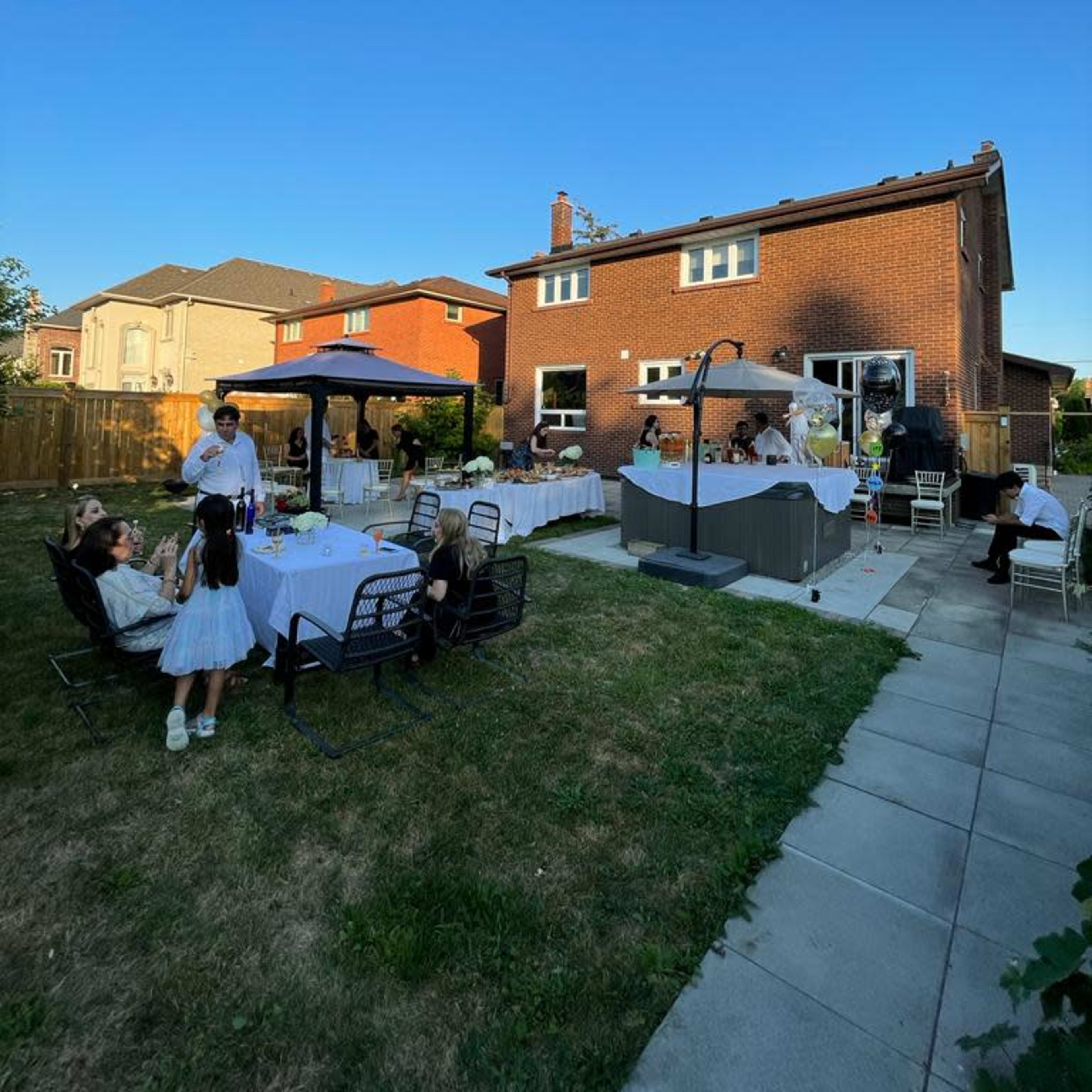 A backyard gathering features tables set for dining, a gazebo, and guests socializing near a house with a clear blue sky above.