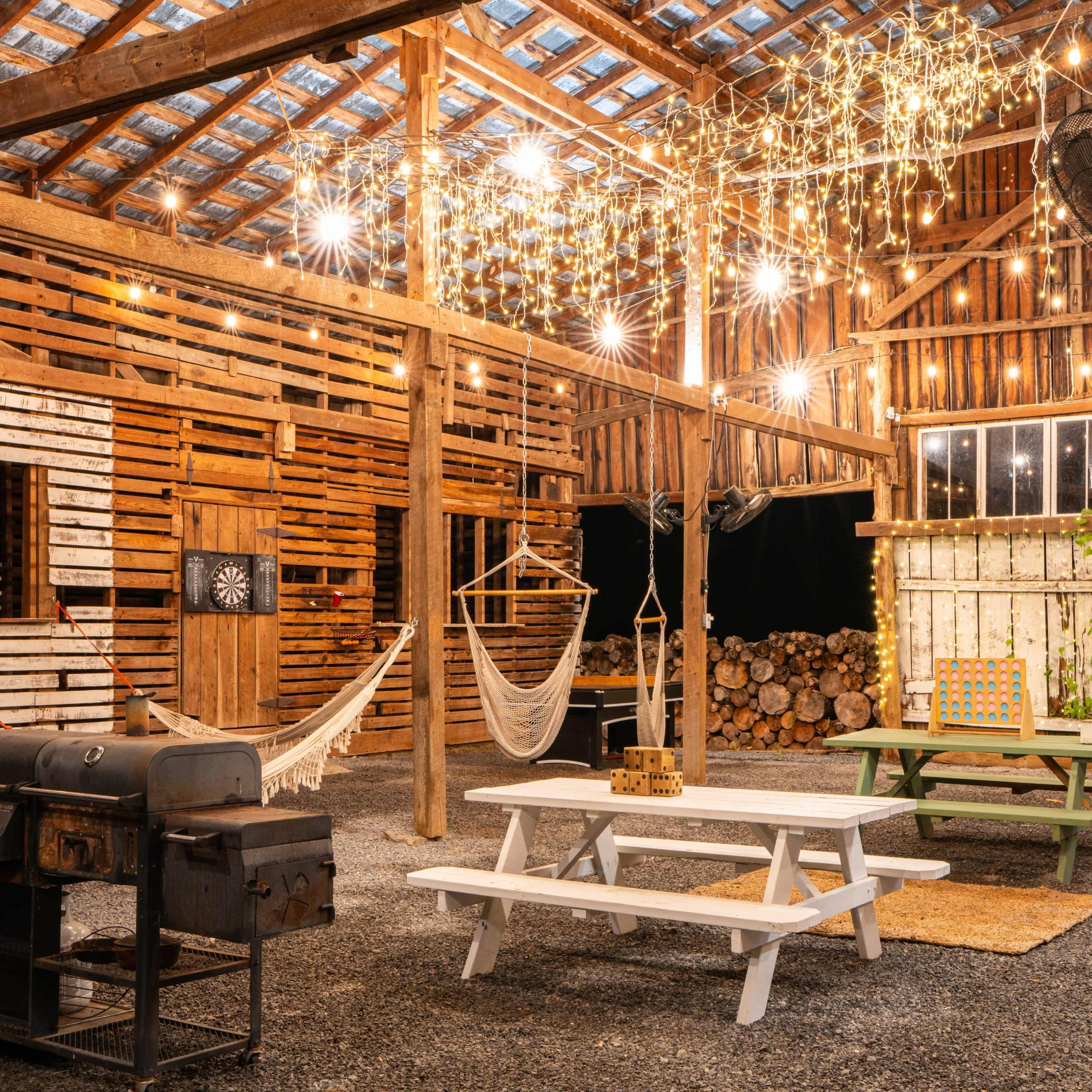 The scene features a rustic barn interior with wooden structures, string lights overhead, a picnic table, a grill, and hammocks hanging between the beams.