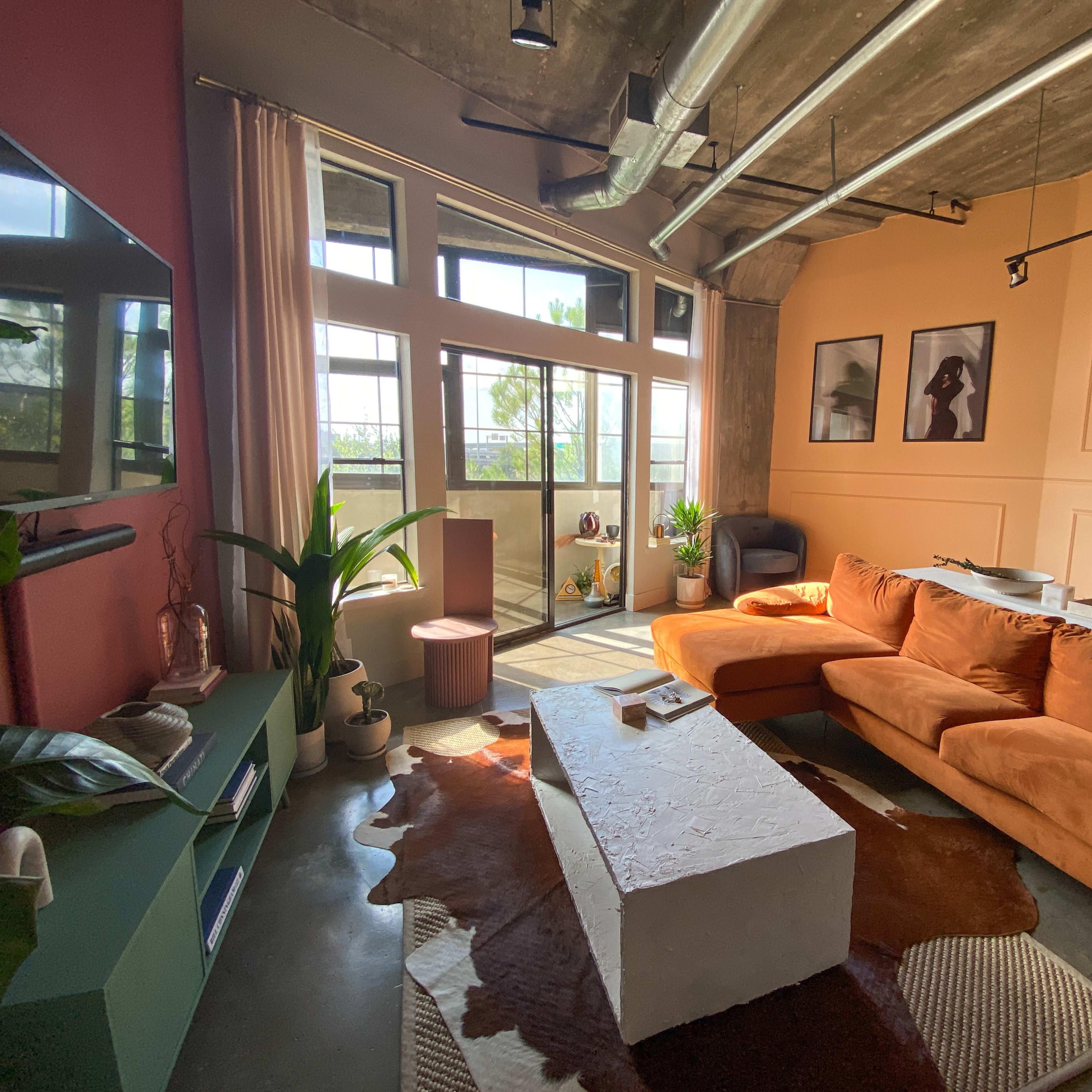 A modern living room with a large orange sectional sofa, a white coffee table, plants, and large windows that let in natural light.