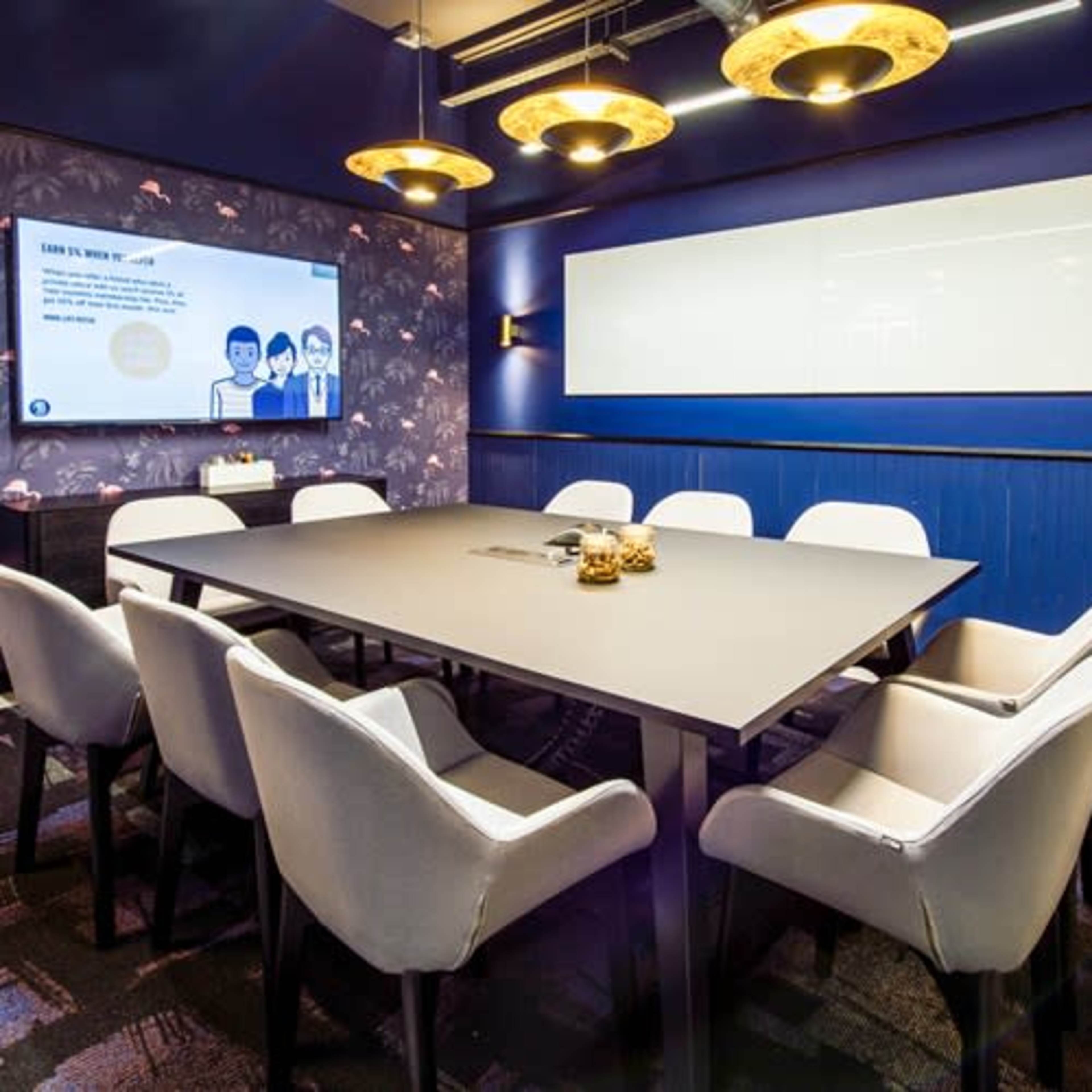 A conference room features a large rectangular table surrounded by upholstered chairs, with a projector screen and decorative lighting on the walls.