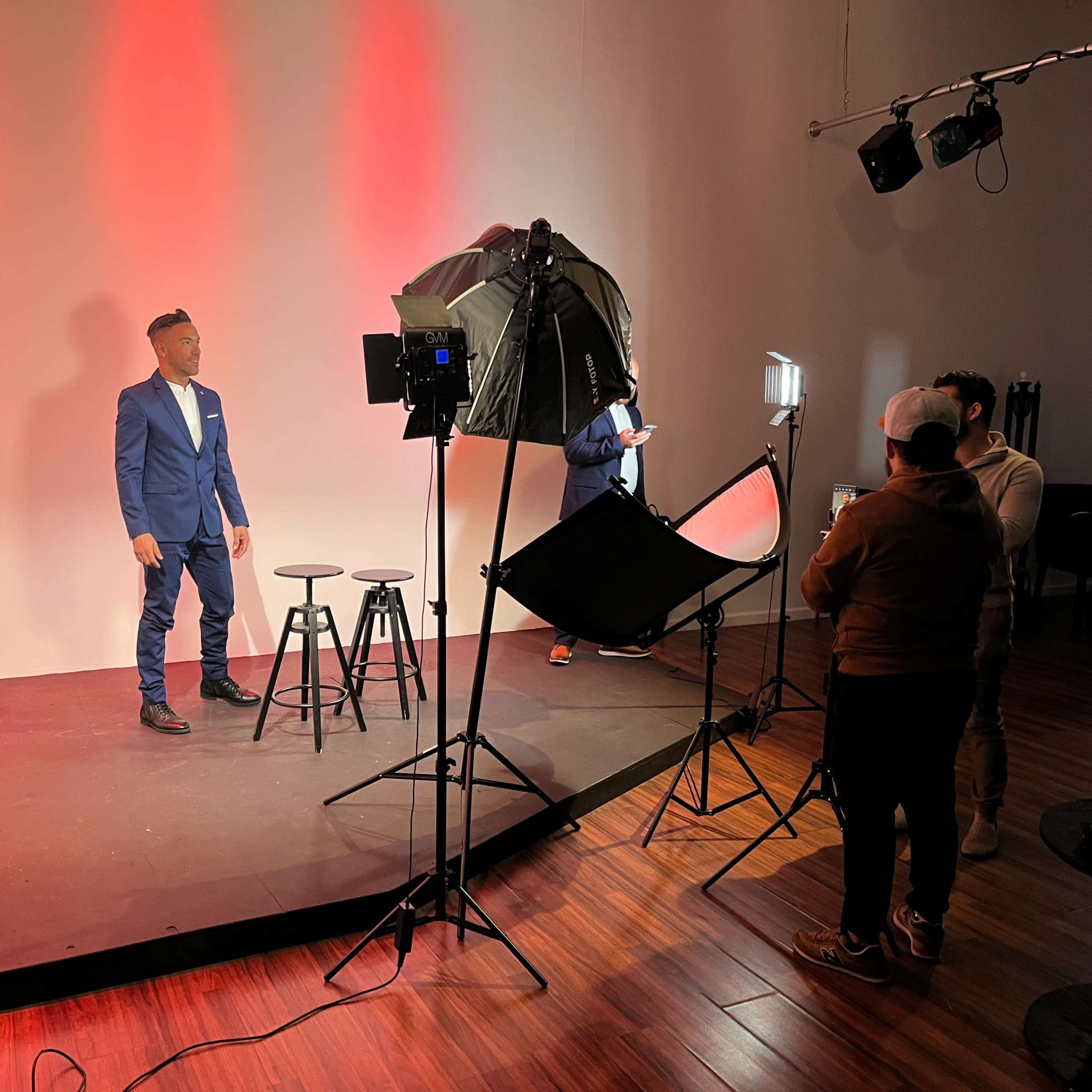 A man in a blue suit stands on a stage with two black stools, while a crew sets up lighting equipment in the background.