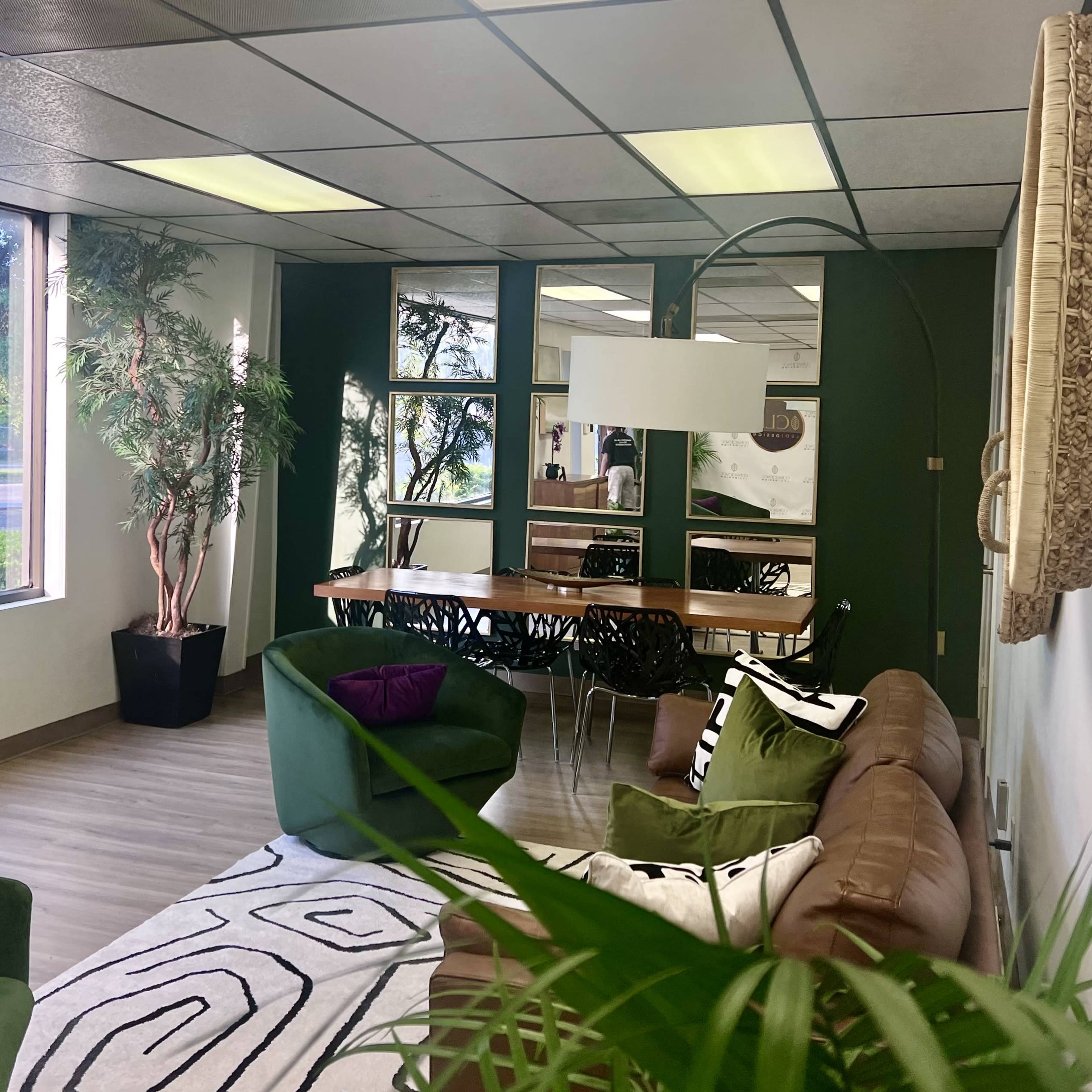 A modern interior with green walls, a wooden dining table, a large light fixture, lounge seating, and houseplants.