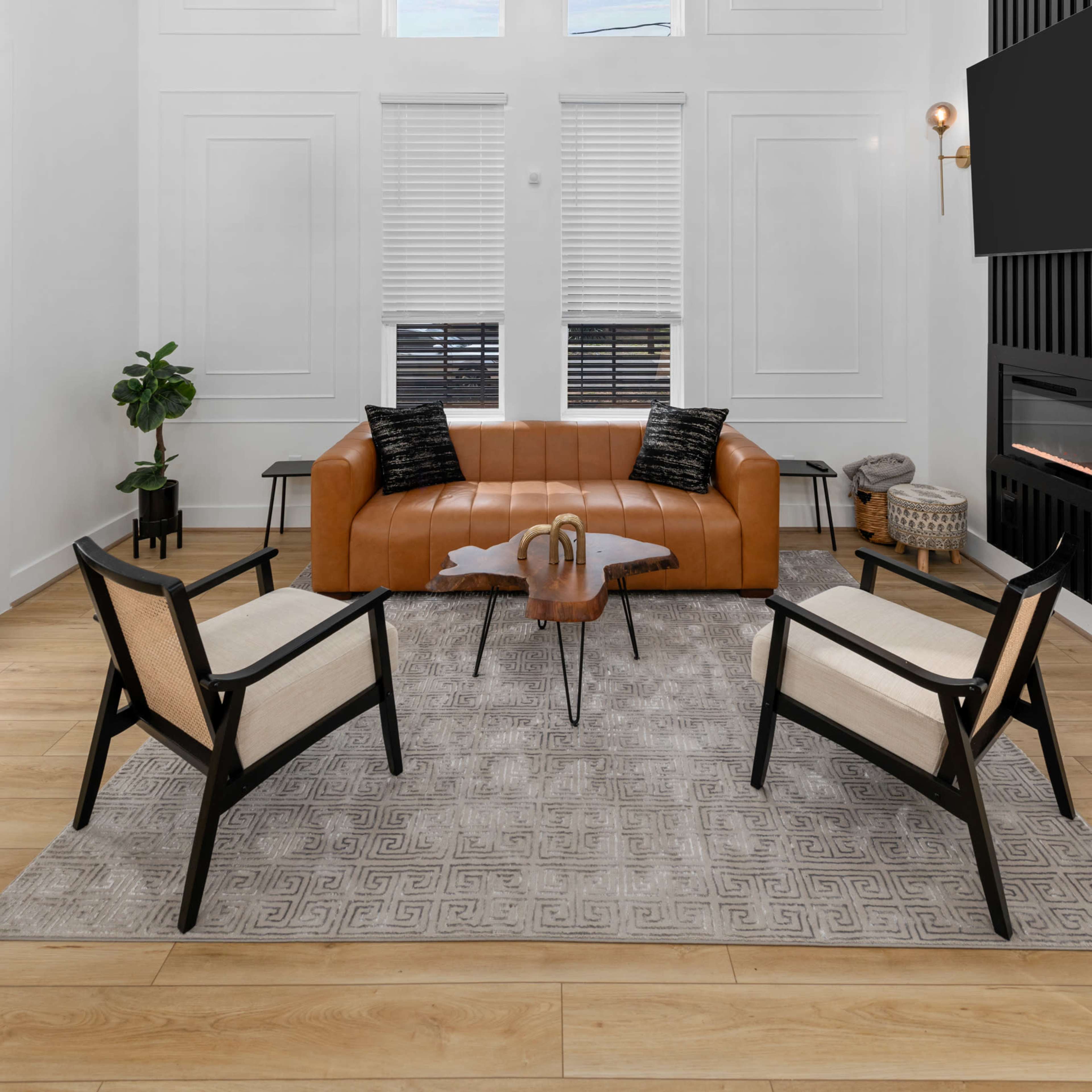 The living room features a high ceiling, large windows, a brown leather sofa, and two black and beige chairs surrounding a central glass coffee table.