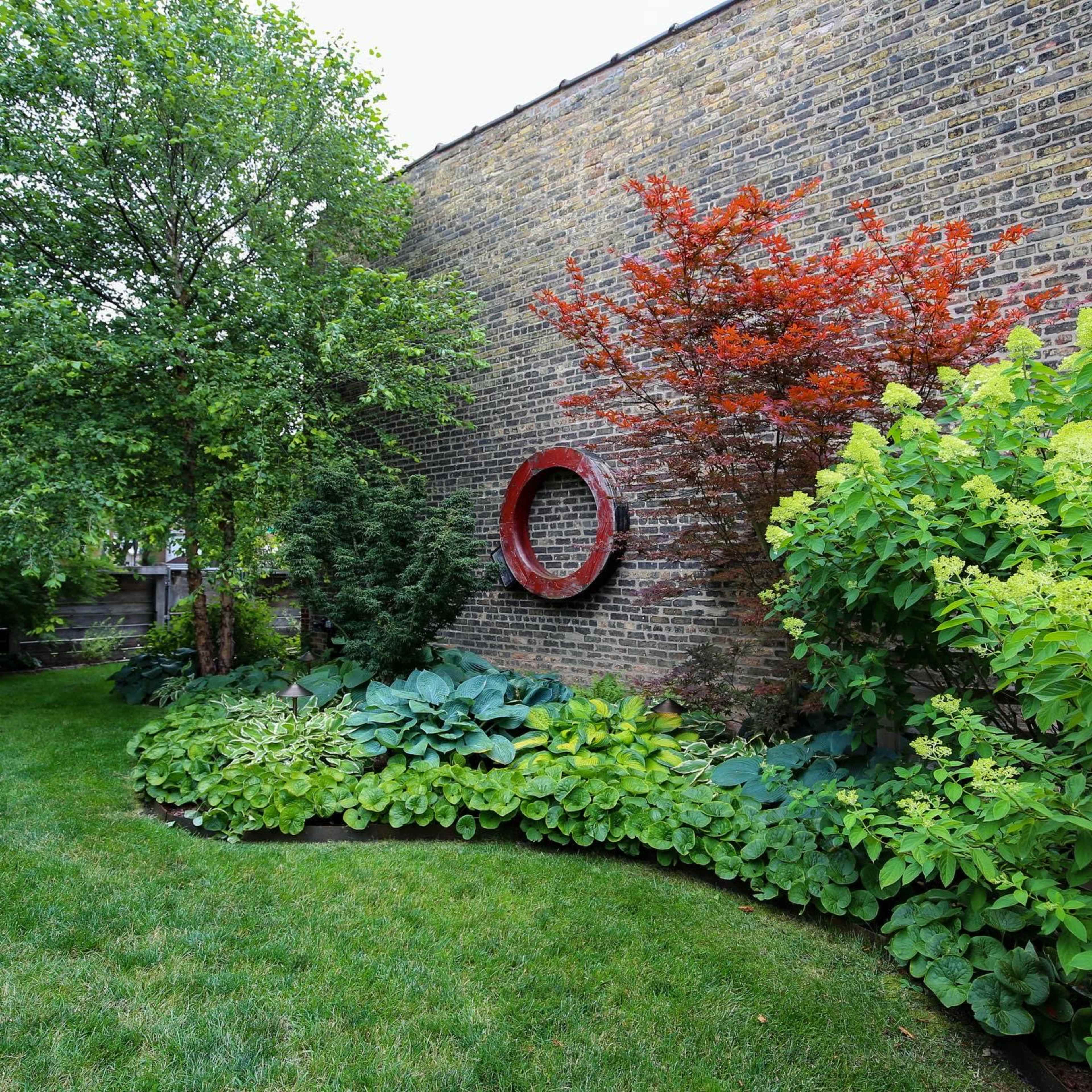 A garden with a curved path lined with various plants, a red circular sculpture on a brick wall, and trees providing shade.