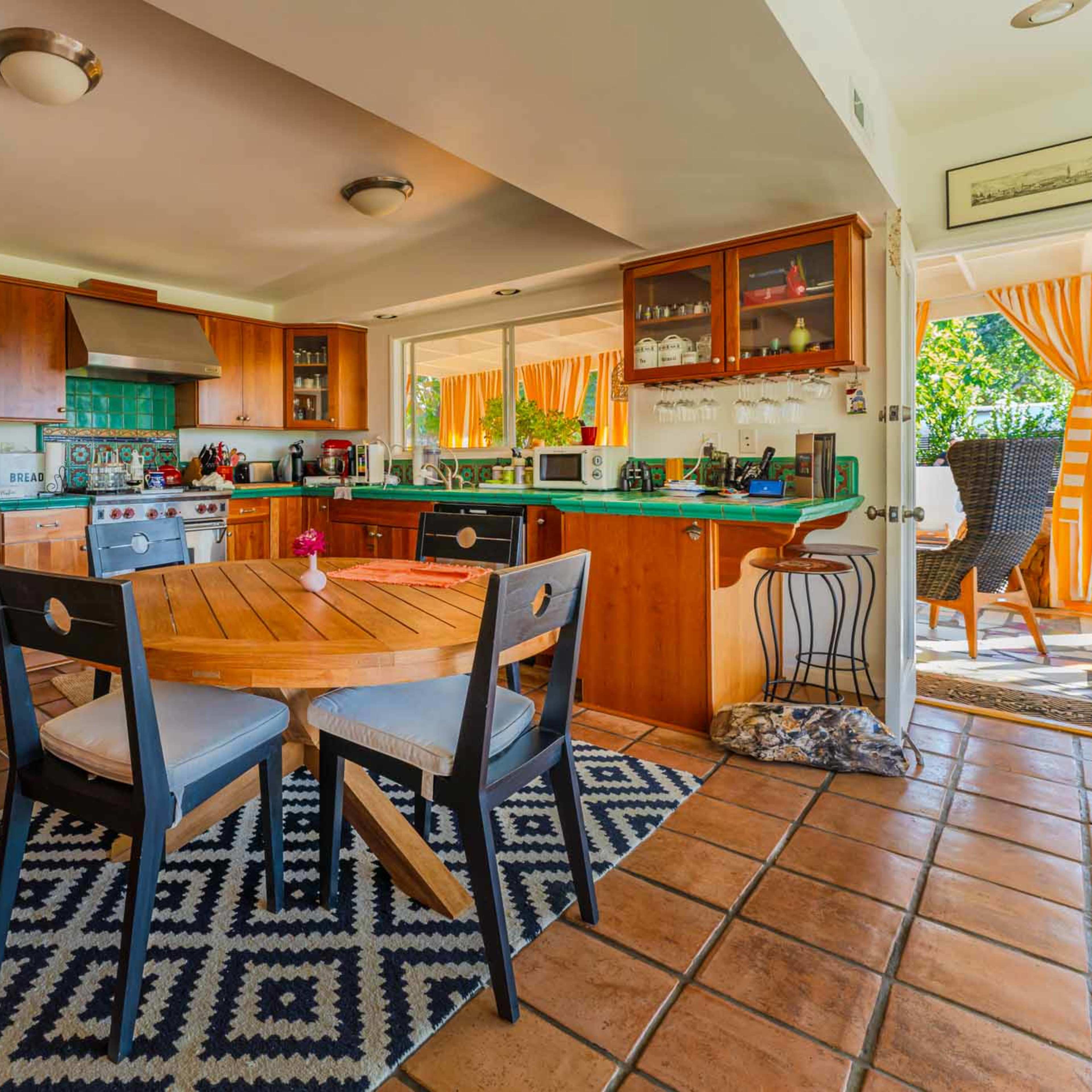 A kitchen with wooden cabinets, a round dining table, and tile flooring features large windows letting in natural light.