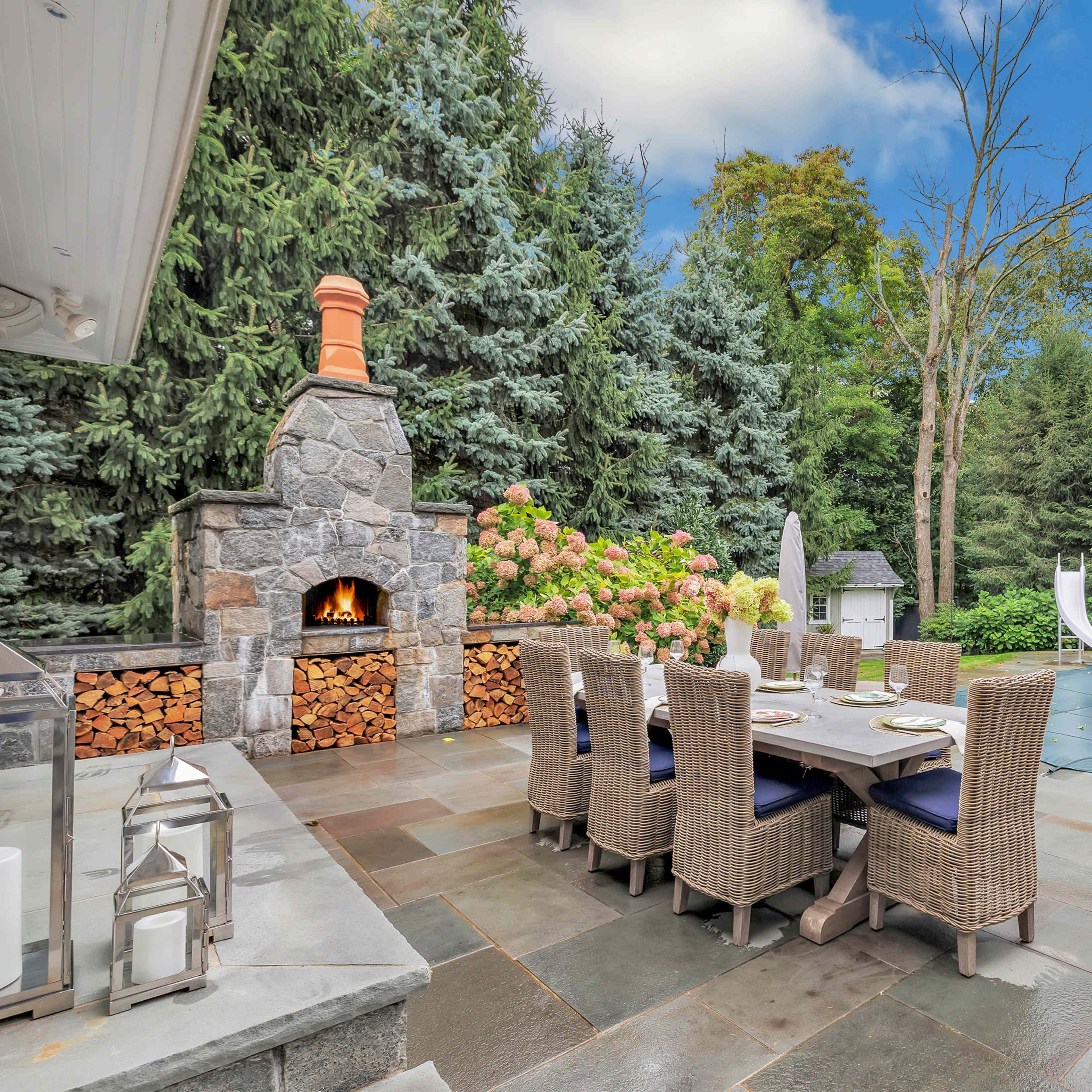 A stone patio features a dining table set for eight, with a fireplace and a swimming pool equipped with a slide in the background, surrounded by tall trees.
