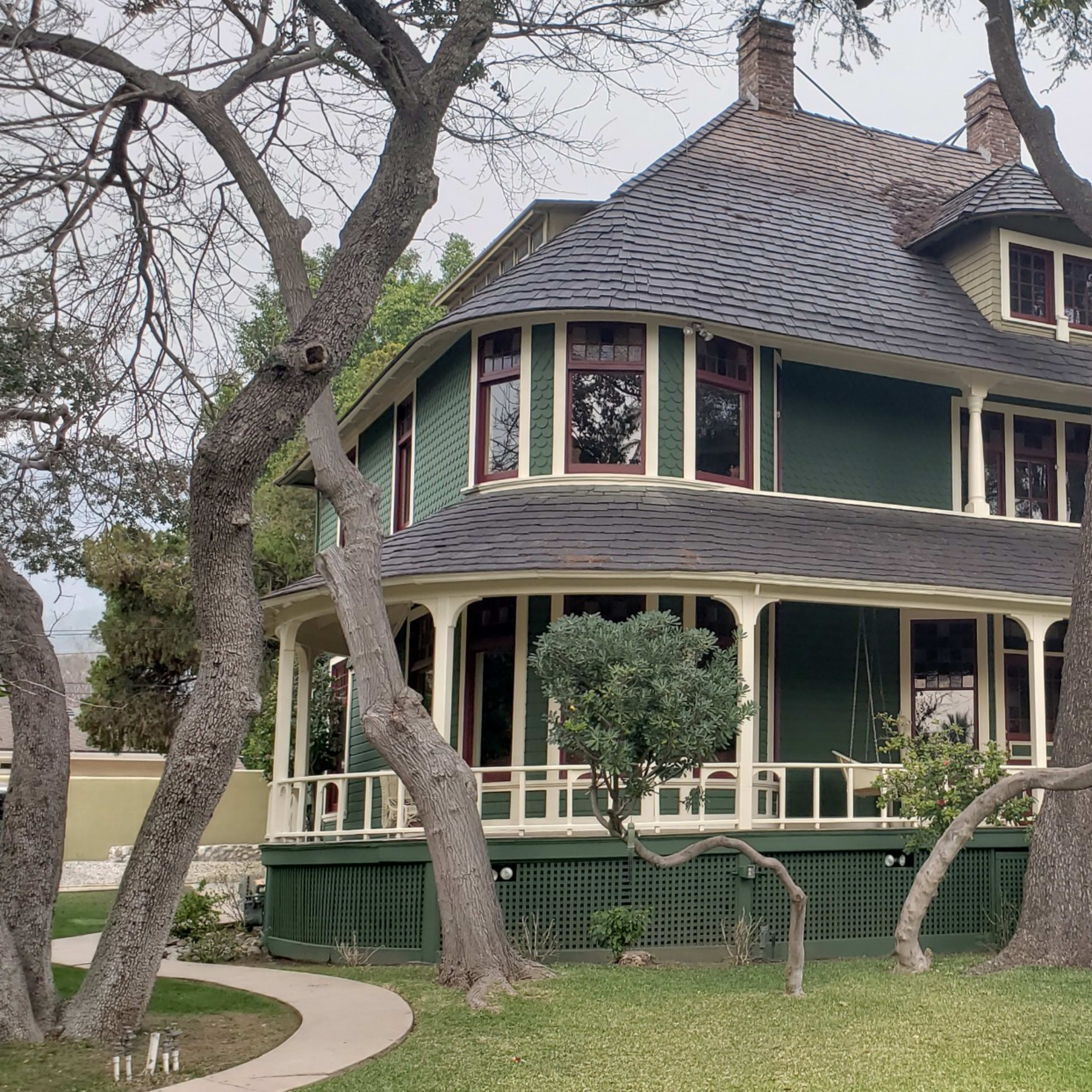 A large green Victorian-style house with a wraparound porch, surrounded by trees and landscaped grass.