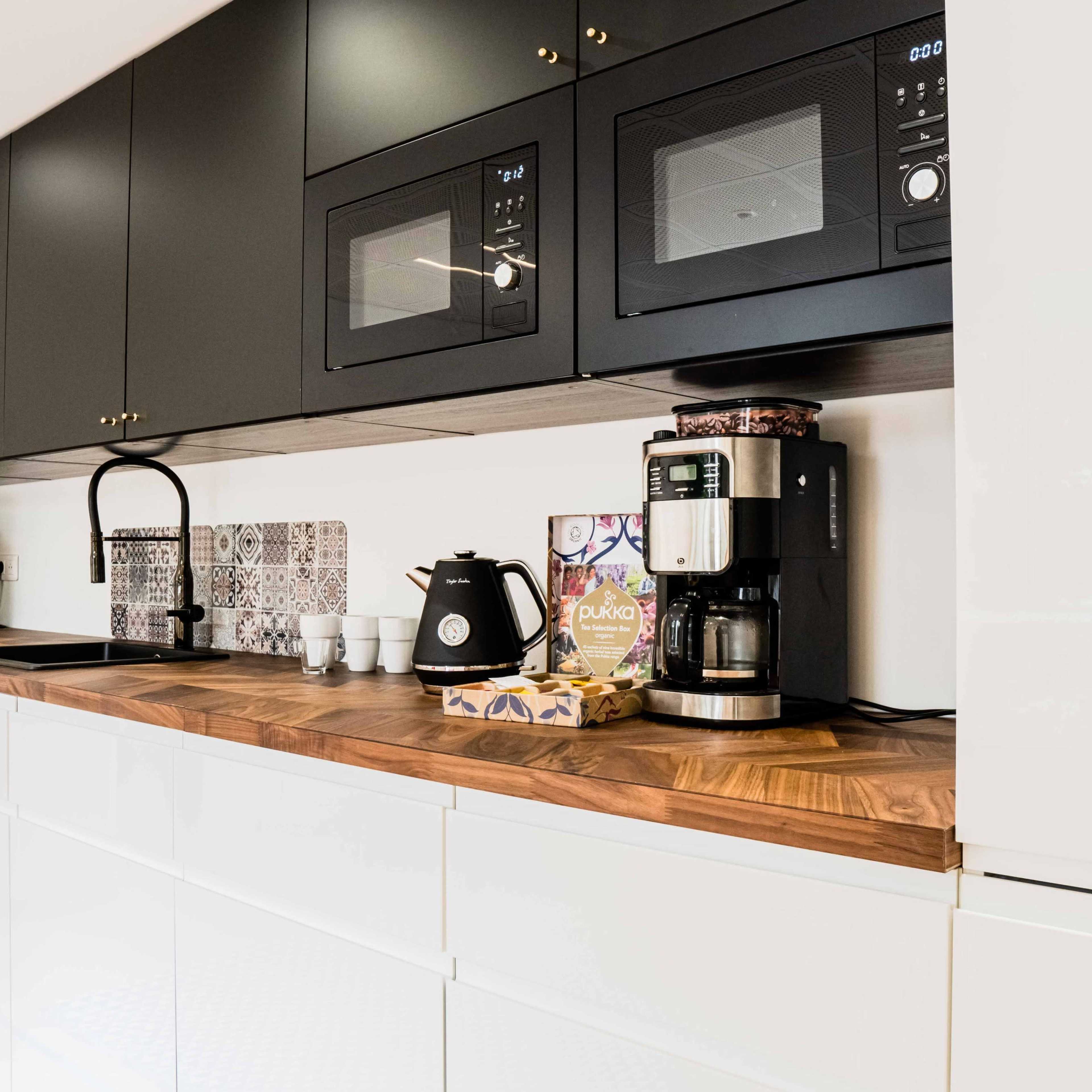 A modern kitchen with black upper cabinets, two microwaves, a coffee maker, and a wooden countertop.