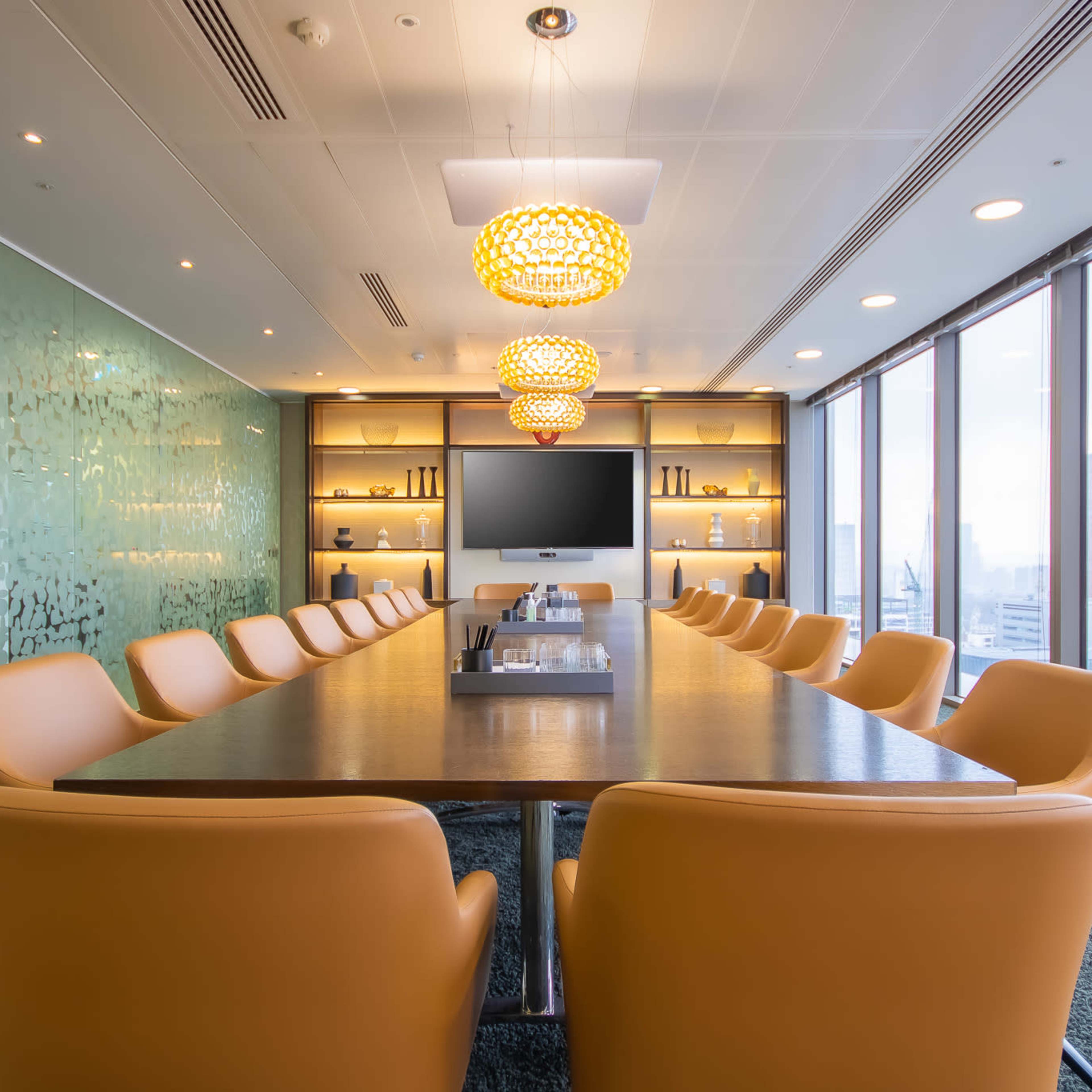 A modern conference room features a long wooden table surrounded by orange chairs, with large windows offering a view of the city and decorative wall elements.