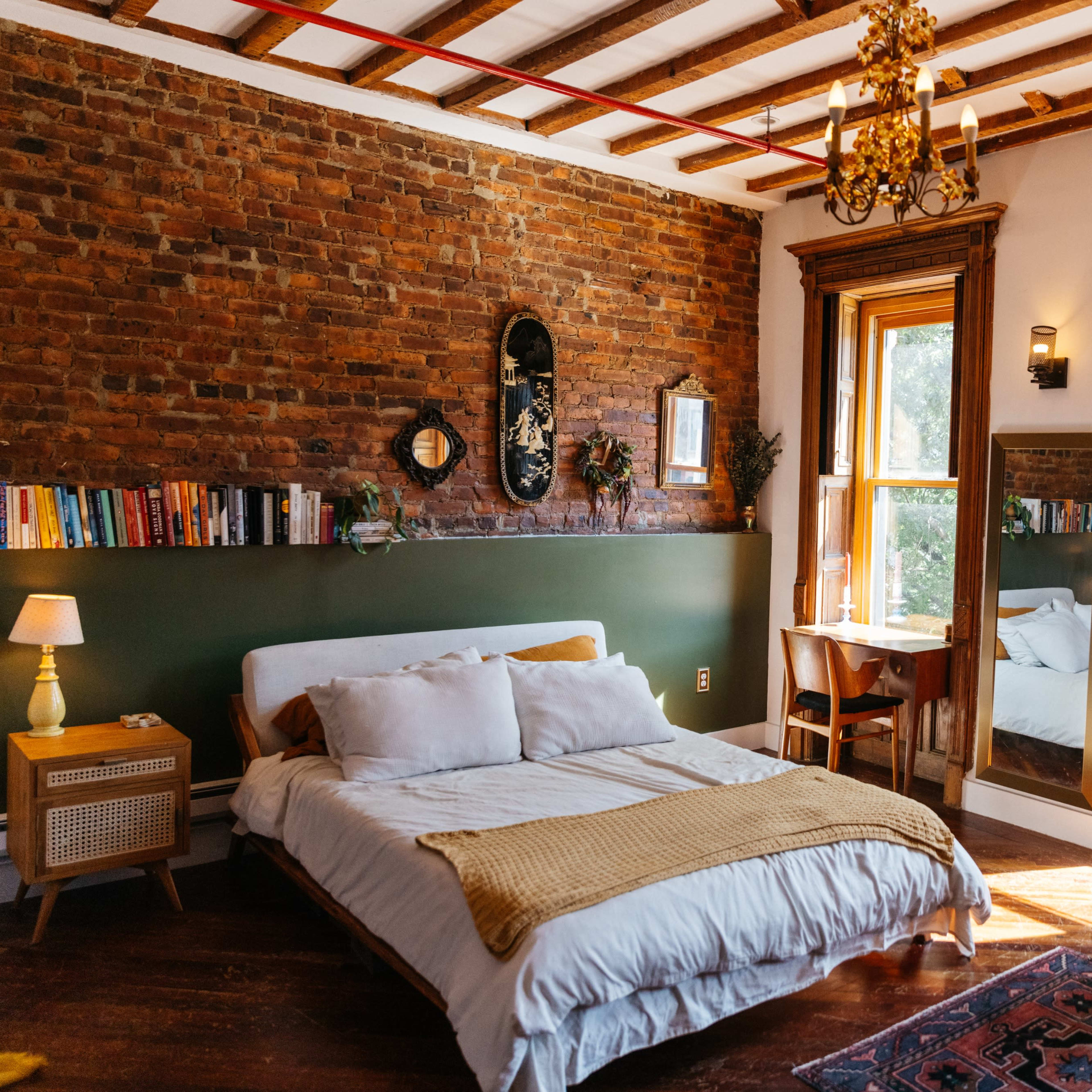 A well-lit bedroom features a mix of exposed brick walls, a bed with white linens, a wooden table, and a cozy seating area.