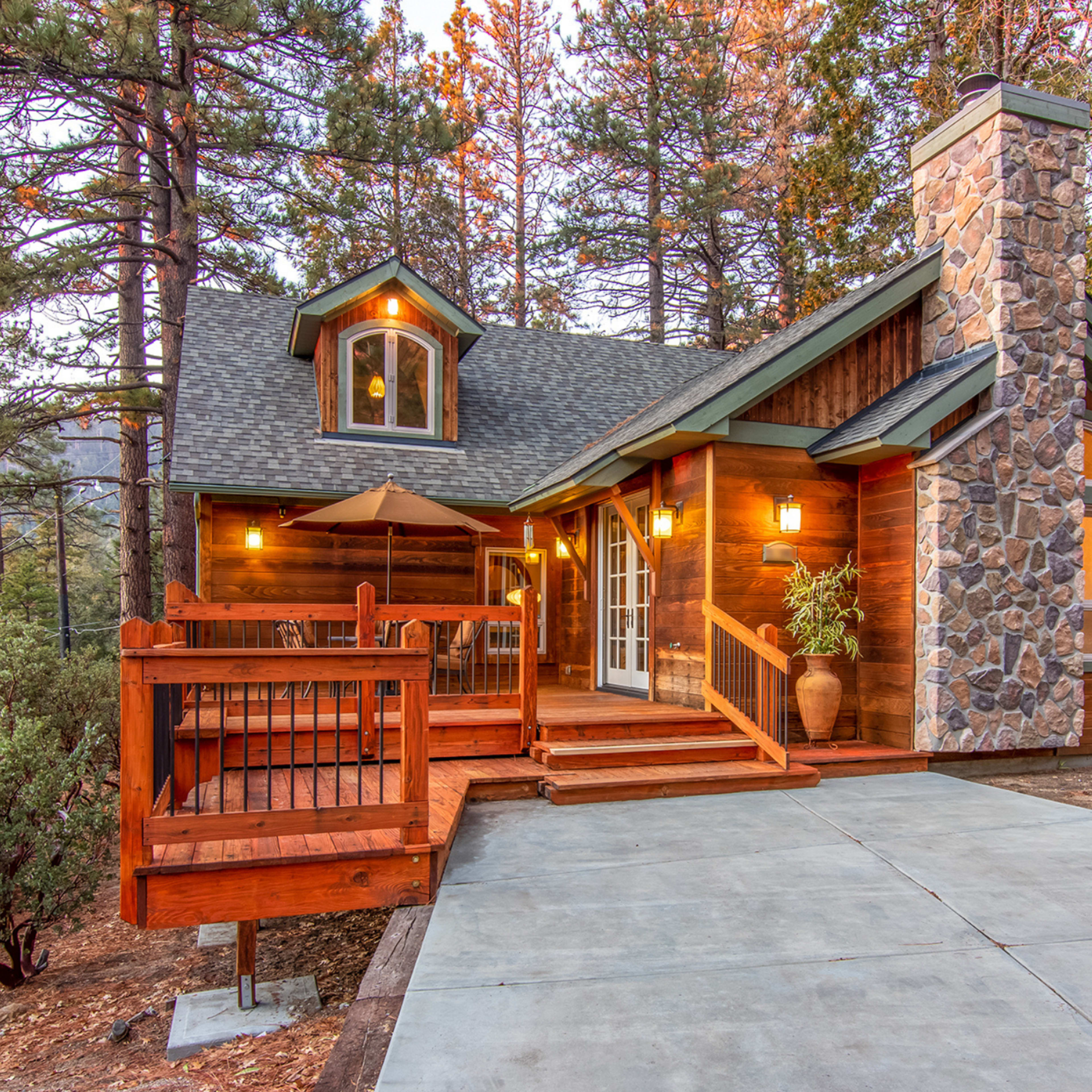 A wooden cabin with a stone chimney features a deck and a concrete driveway, surrounded by trees.