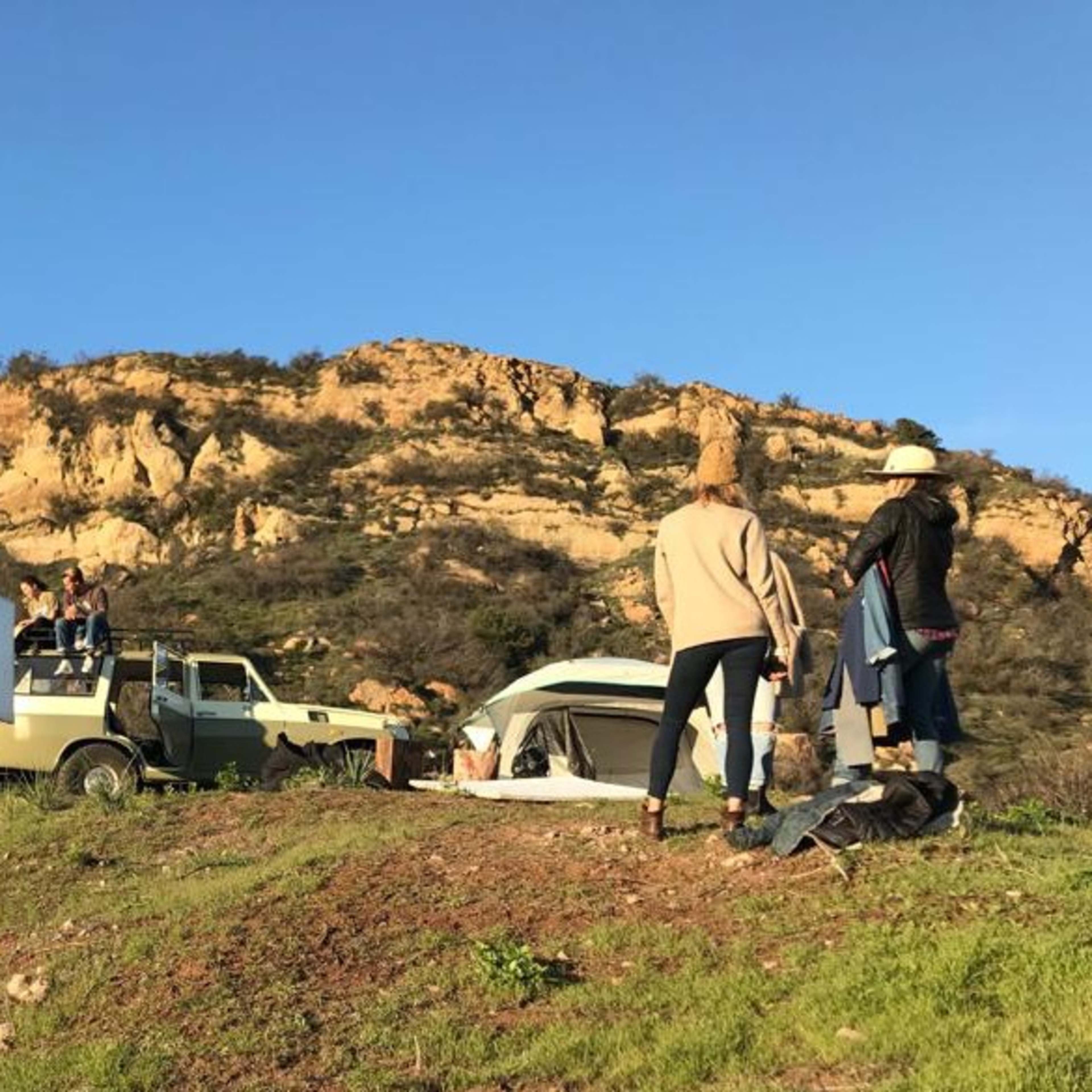 A group of people is filming on a hillside with a vintage truck and a tent near a rocky landscape.