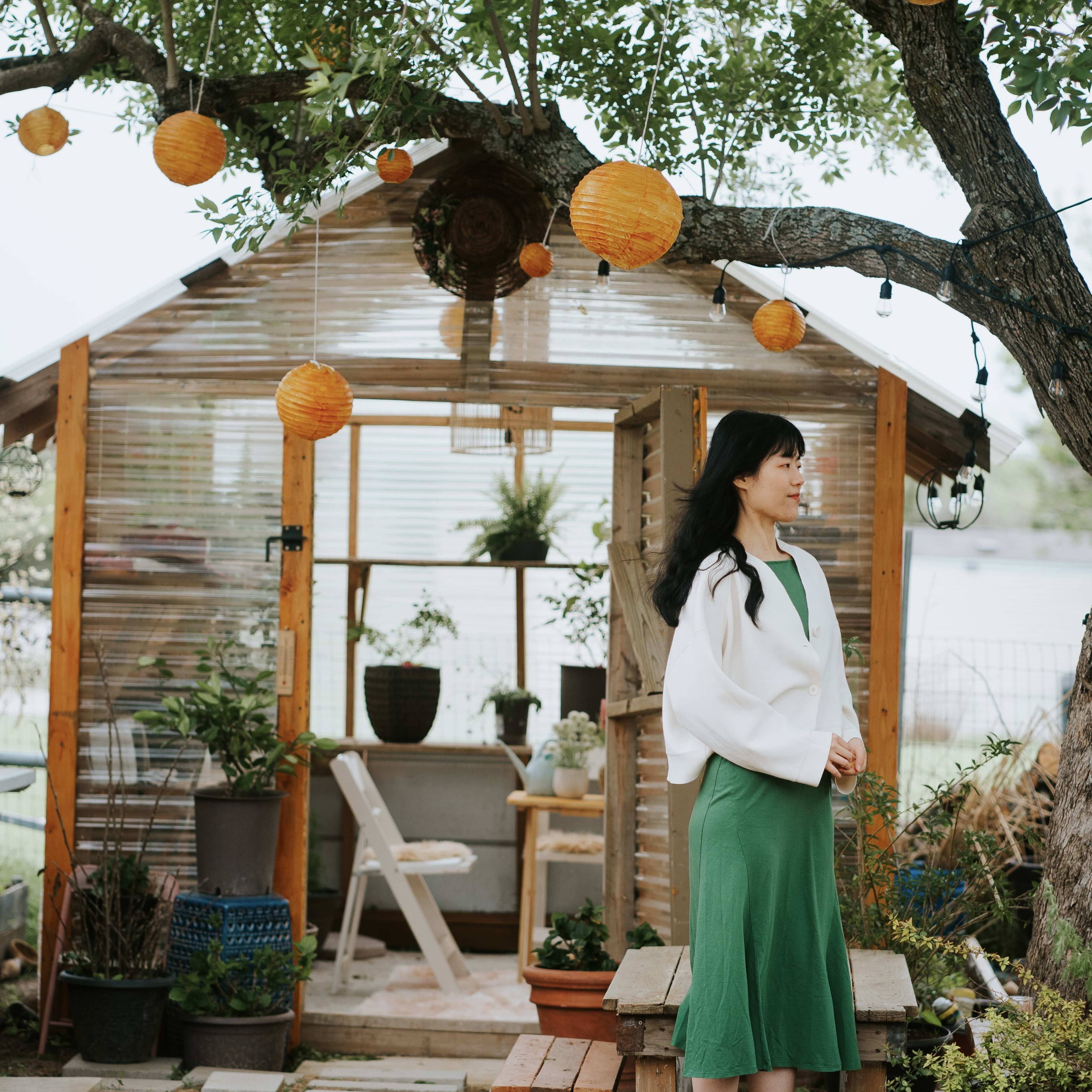 A woman in a green dress stands outside a greenhouse adorned with hanging lanterns and surrounded by potted plants.